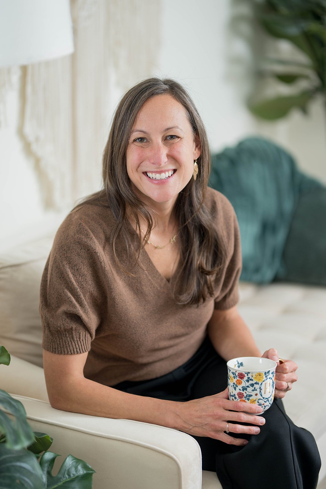 A woman with long brown hair and a black sweater smiling while sitting on a beige chair in a well-lit room with cream-colored curtains and a lamp.