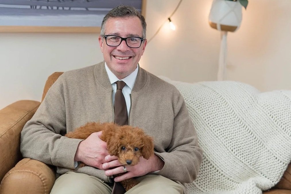 A smiling man sitting on a brown leather couch in a room with two beach-themed black and white framed photographs on the wall behind him.