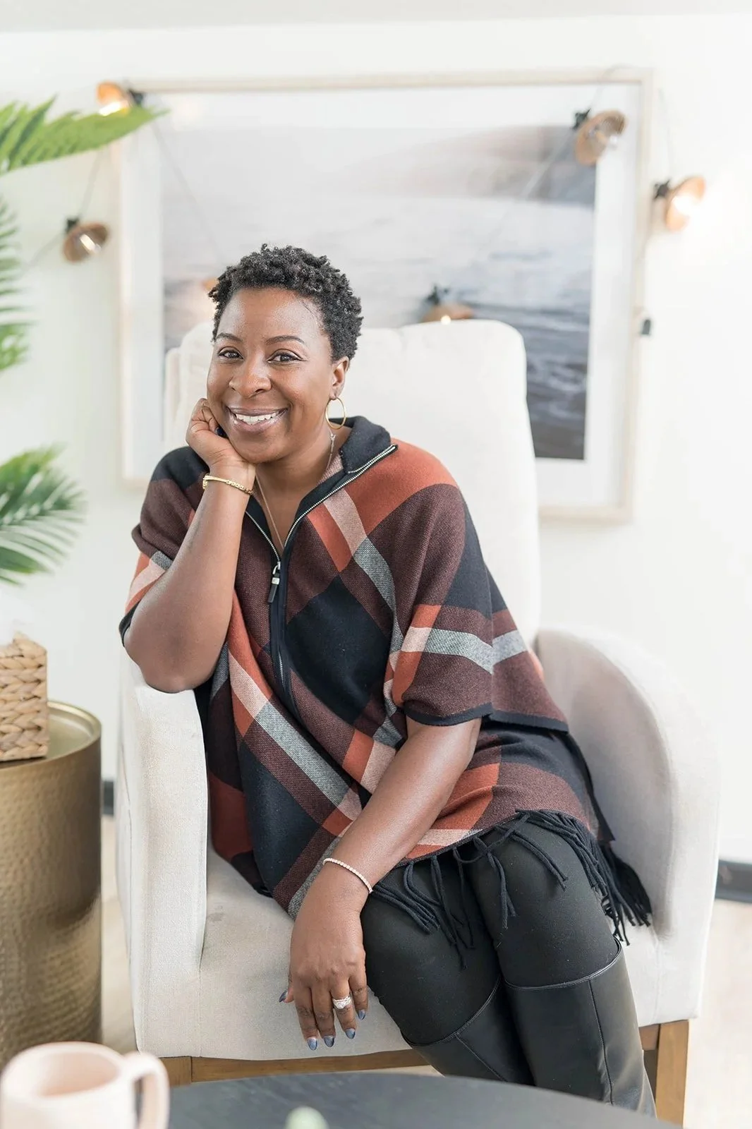 A woman with short curly hair smiling while resting her chin on her hand in a cozy, well-lit room with blue curtains and modern decor.
