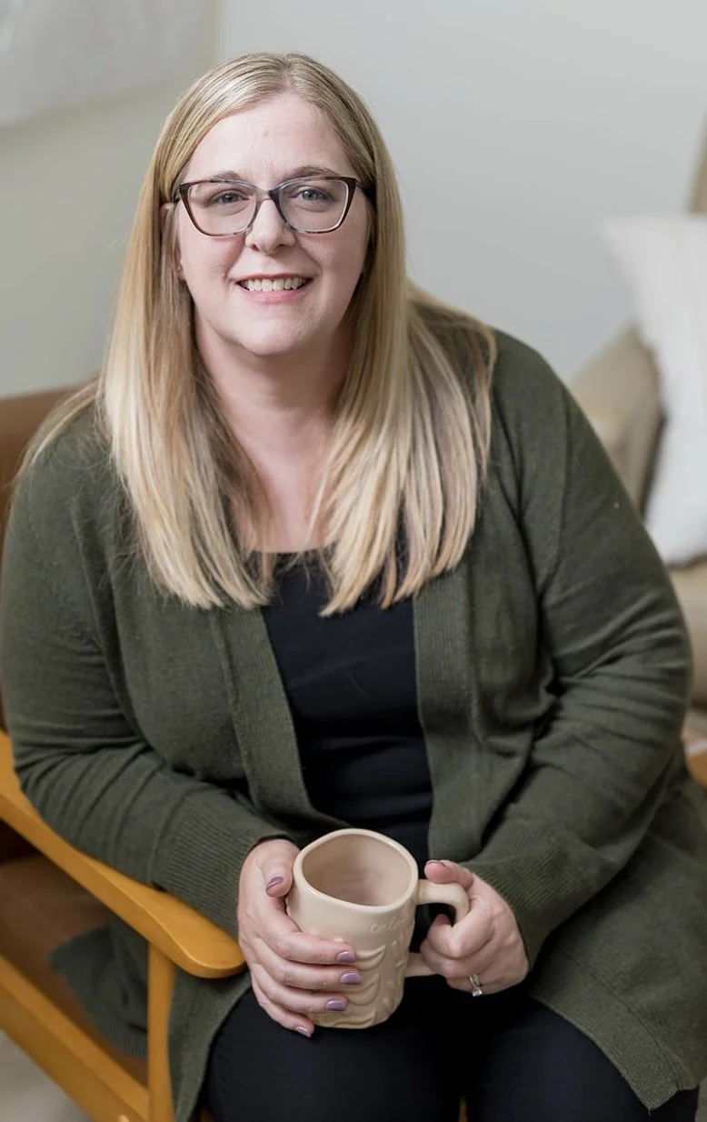 A woman with blonde hair, glasses, and a big smile, sitting on a beige couch with a wooden wall and a plant in the background.