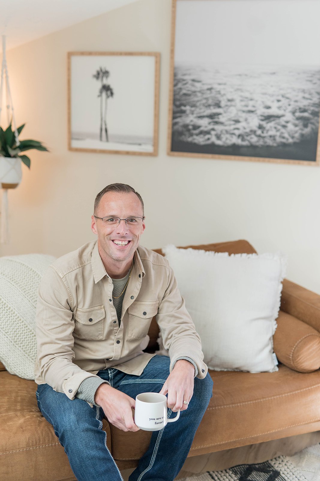 A smiling man wearing glasses and a patterned sweater, sitting indoors with curtains and a window in the background.