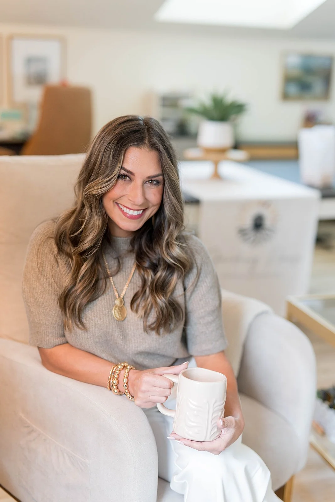 A woman with long, wavy brown hair smiling while sitting on a beige sofa, holding a white mug in her right hand and resting her left hand on her knee, in a cozy living room setting.