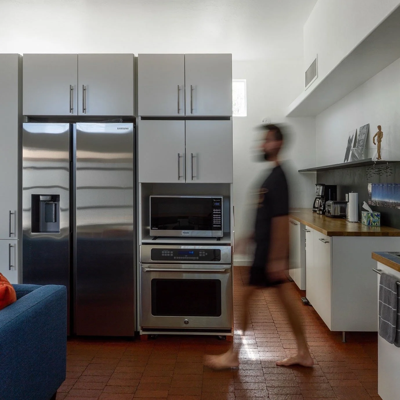 A modern kitchen with stainless steel refrigerator, oven, microwave, and countertop appliances. A person with blurred motion is walking barefoot past the kitchen area.