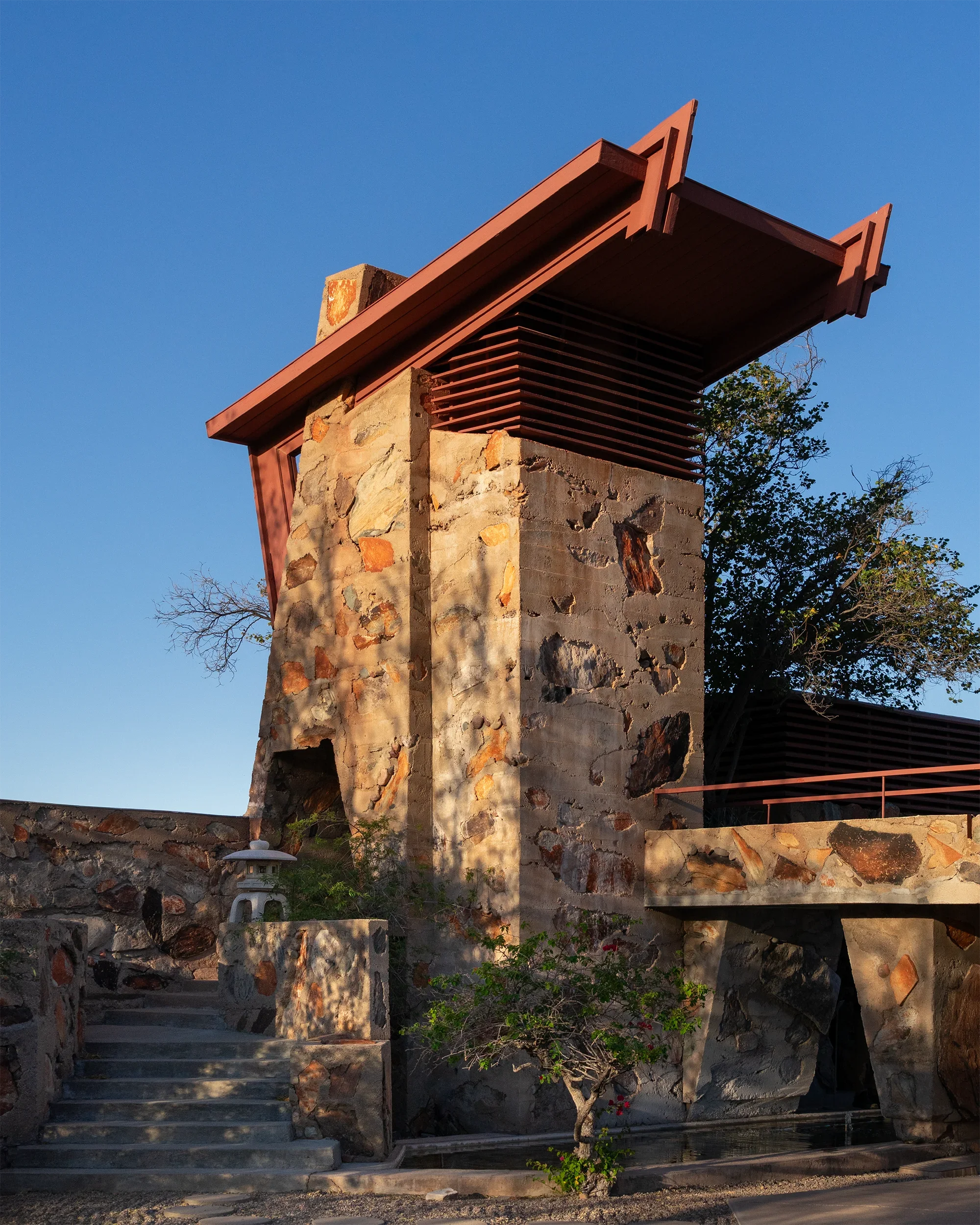 A stone and wood structure with stairs, trees, and a clear blue sky in the background, resembling a Japanese garden or temple setting.