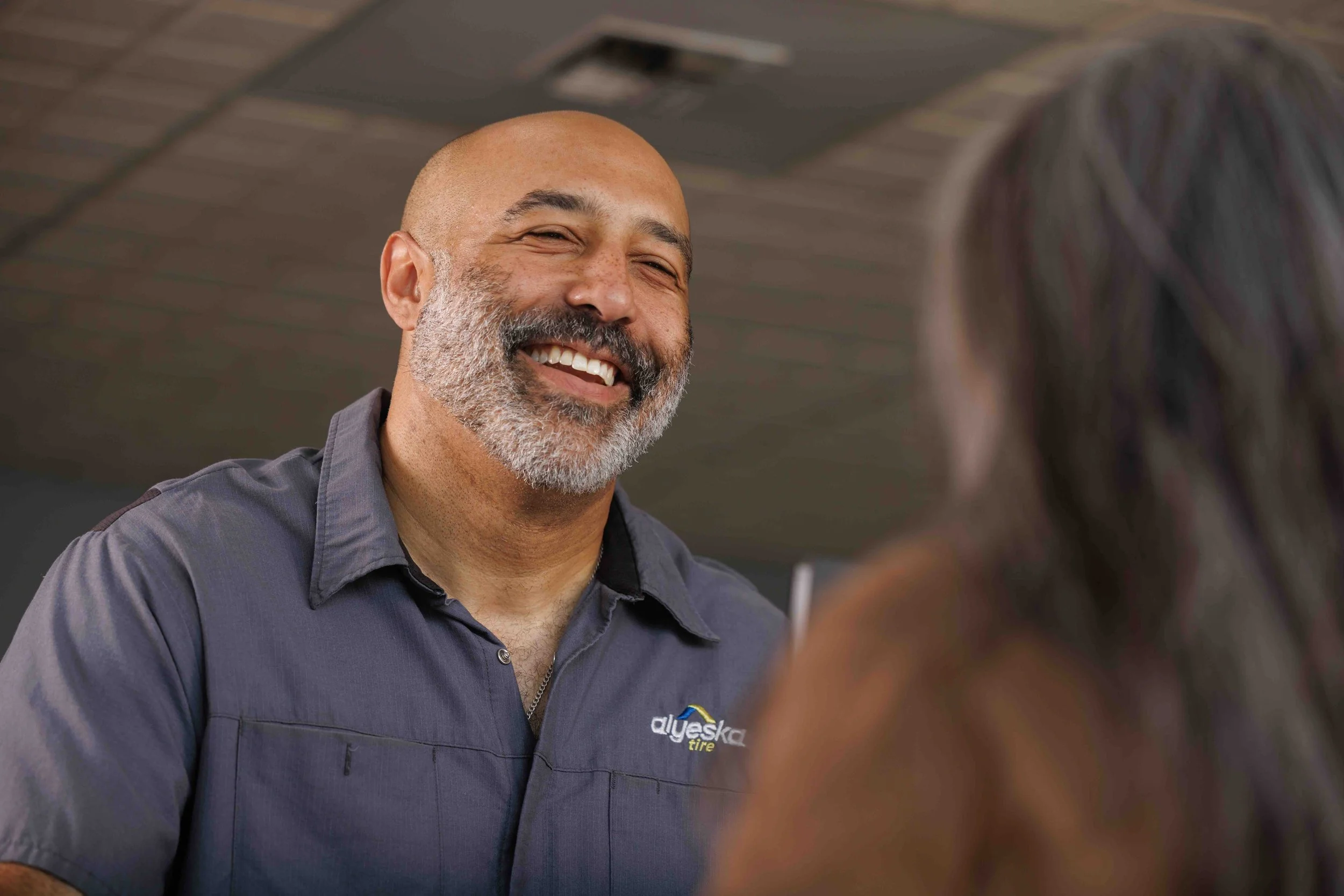 A smiling auto Office Support Specialist in a gray uniform speaking with a customer inside Alyeska Tire.