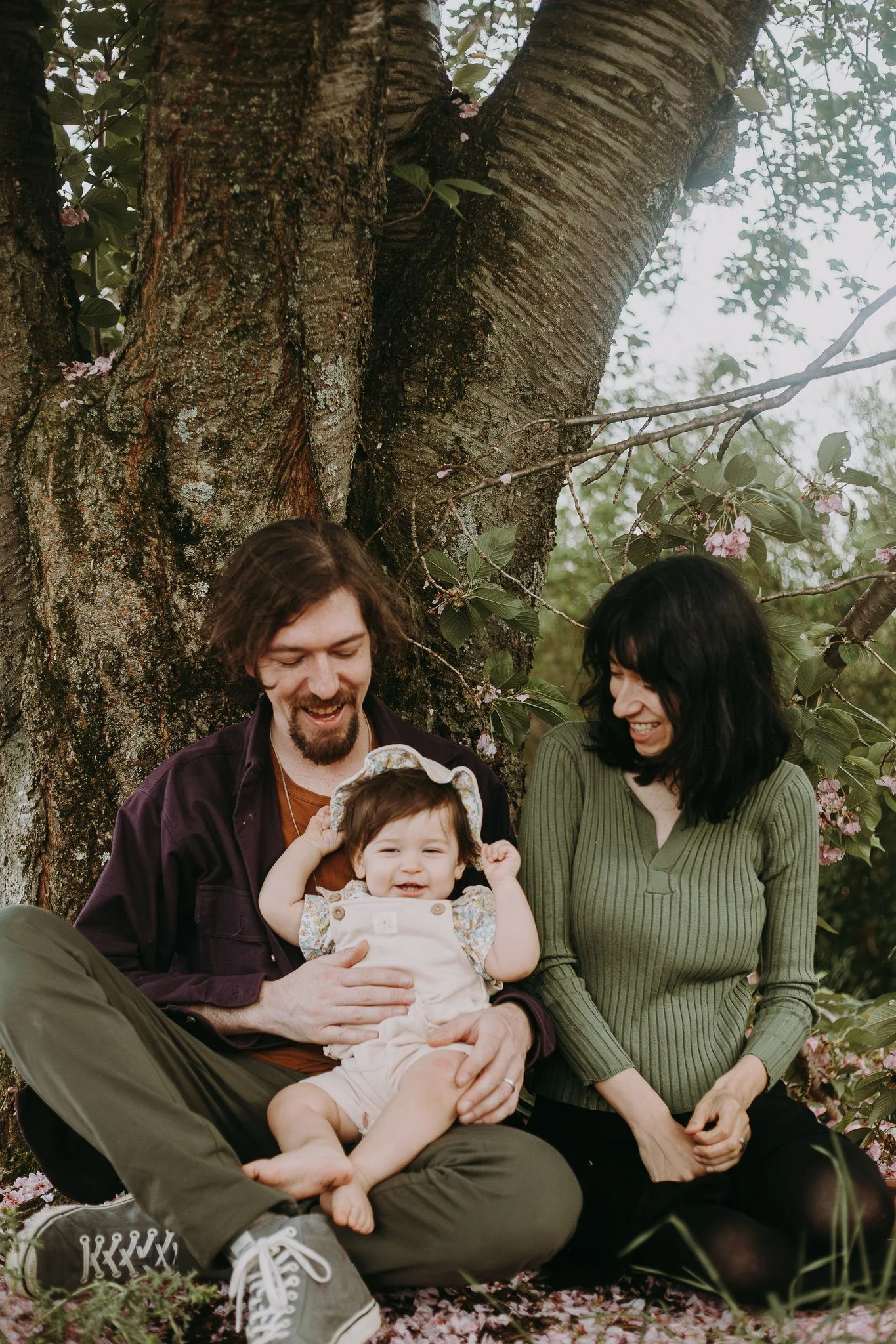 Parents sitting down against a tree smiling down at a baby in their lap