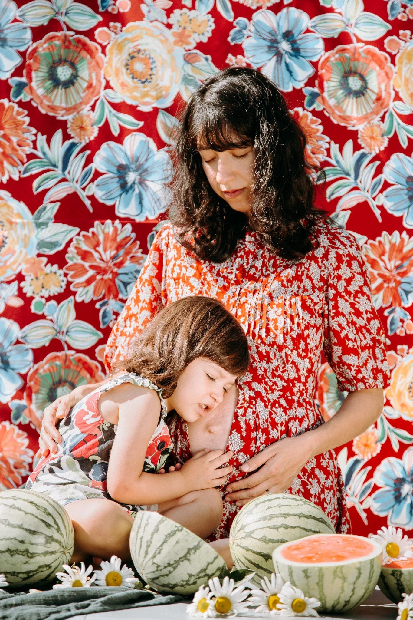 A parent sitting on the ground with their child as they look at watermelons 