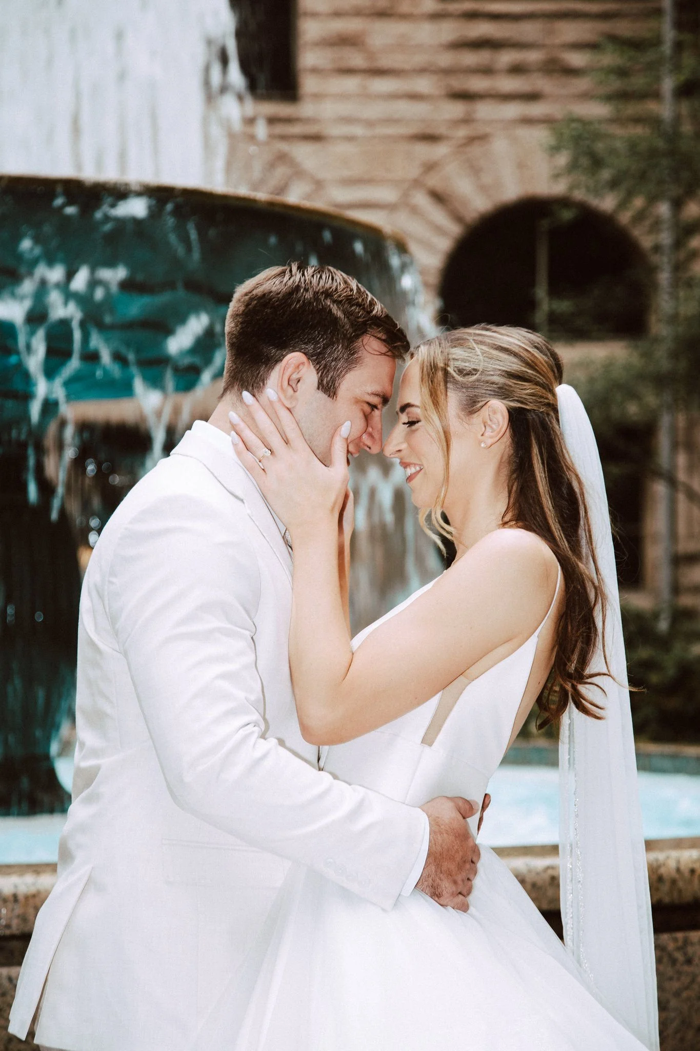 A bride and groom sharing a close, loving moment, smiling with their foreheads touching in front of a waterfall feature.