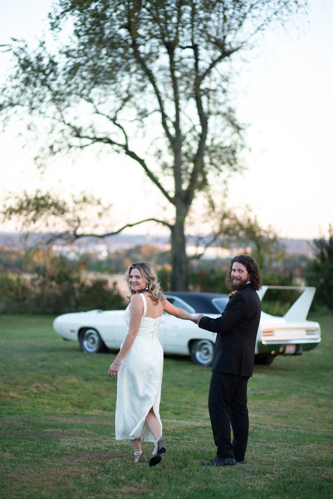 A woman in a white dress and a man in a black suit holding hands, smiling, with a classic white car in the background outdoors during sunset.