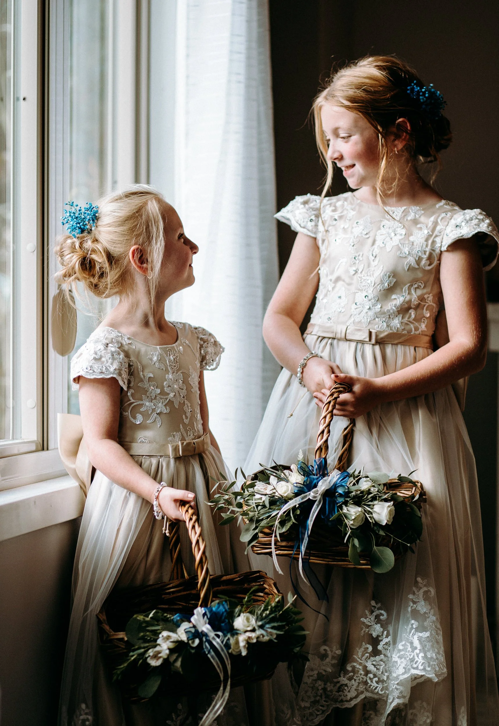 Two young girls in cream-colored lace dresses standing by a window, holding baskets decorated with flowers and ribbons, sharing a moment together.