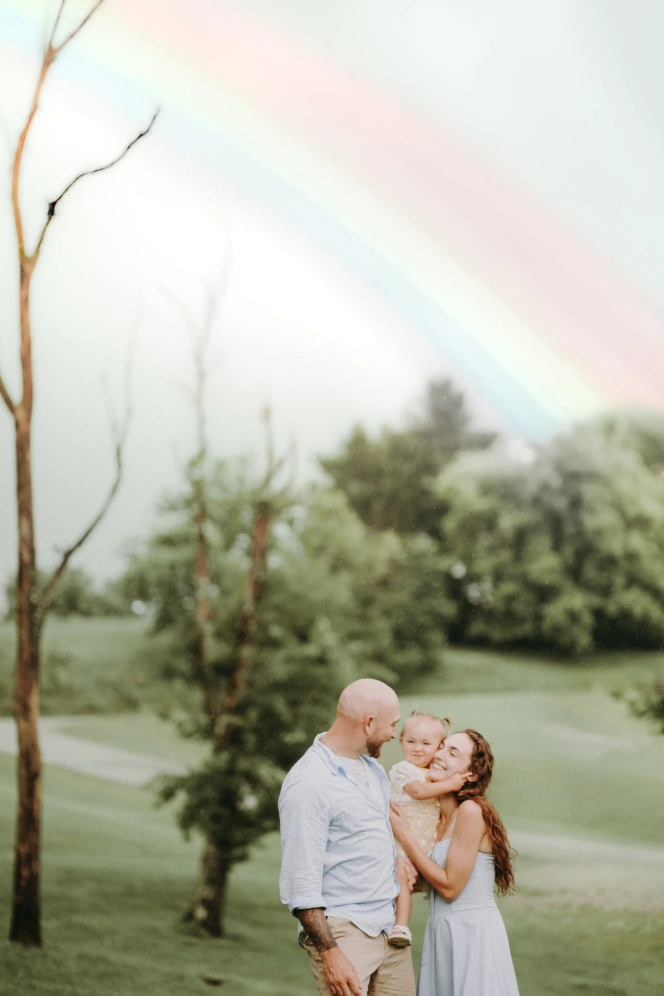 Two parents standing in a field smiling at their daughter in their arms with a rainbow in the background