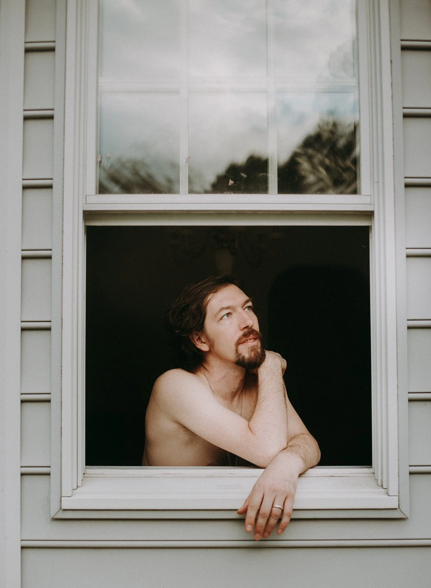 A shirtless man with long hair and a beard resting his chin on his hand at an open window of a house.