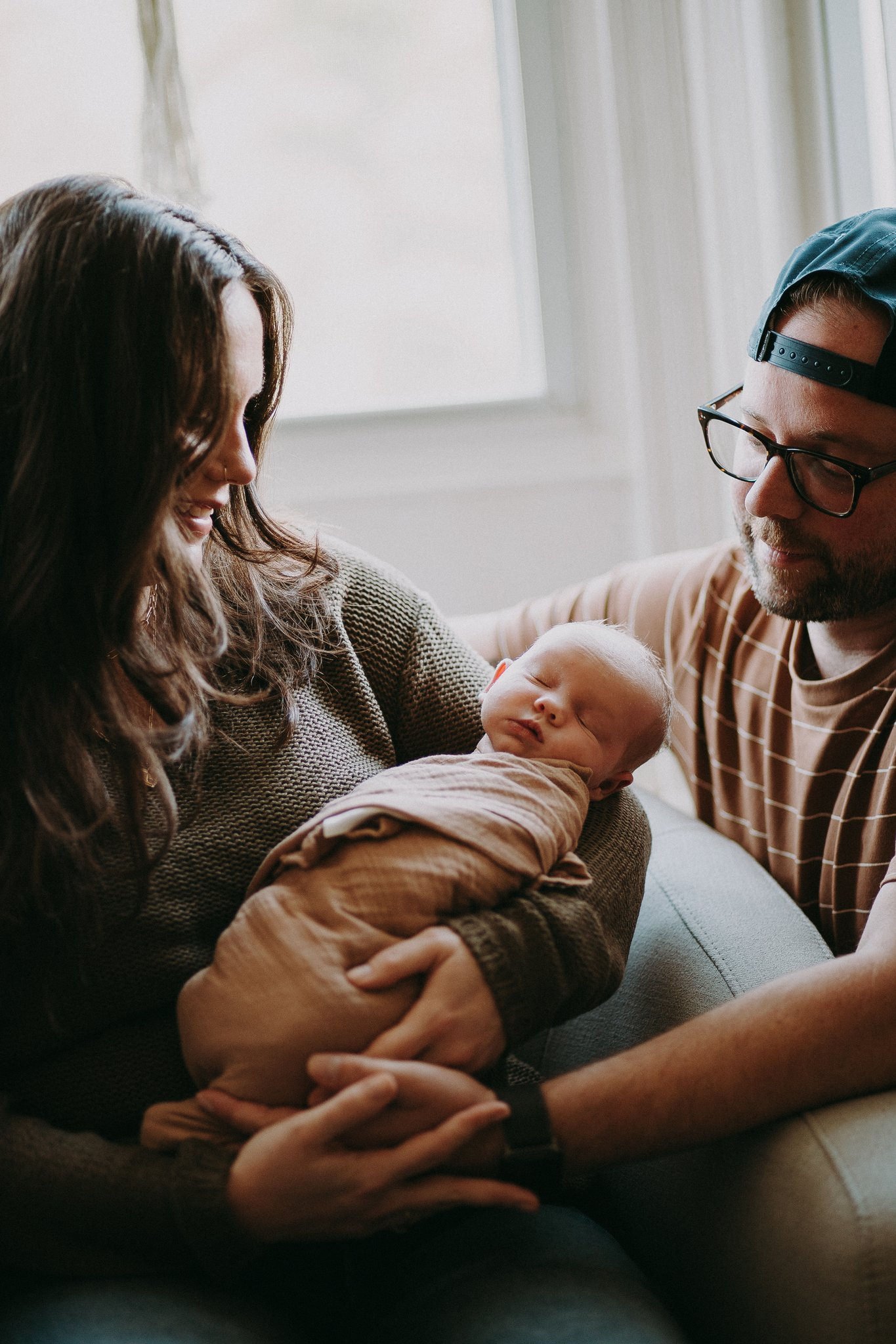 A parent sitting in a chair holding a newborn as their partner is next to them 