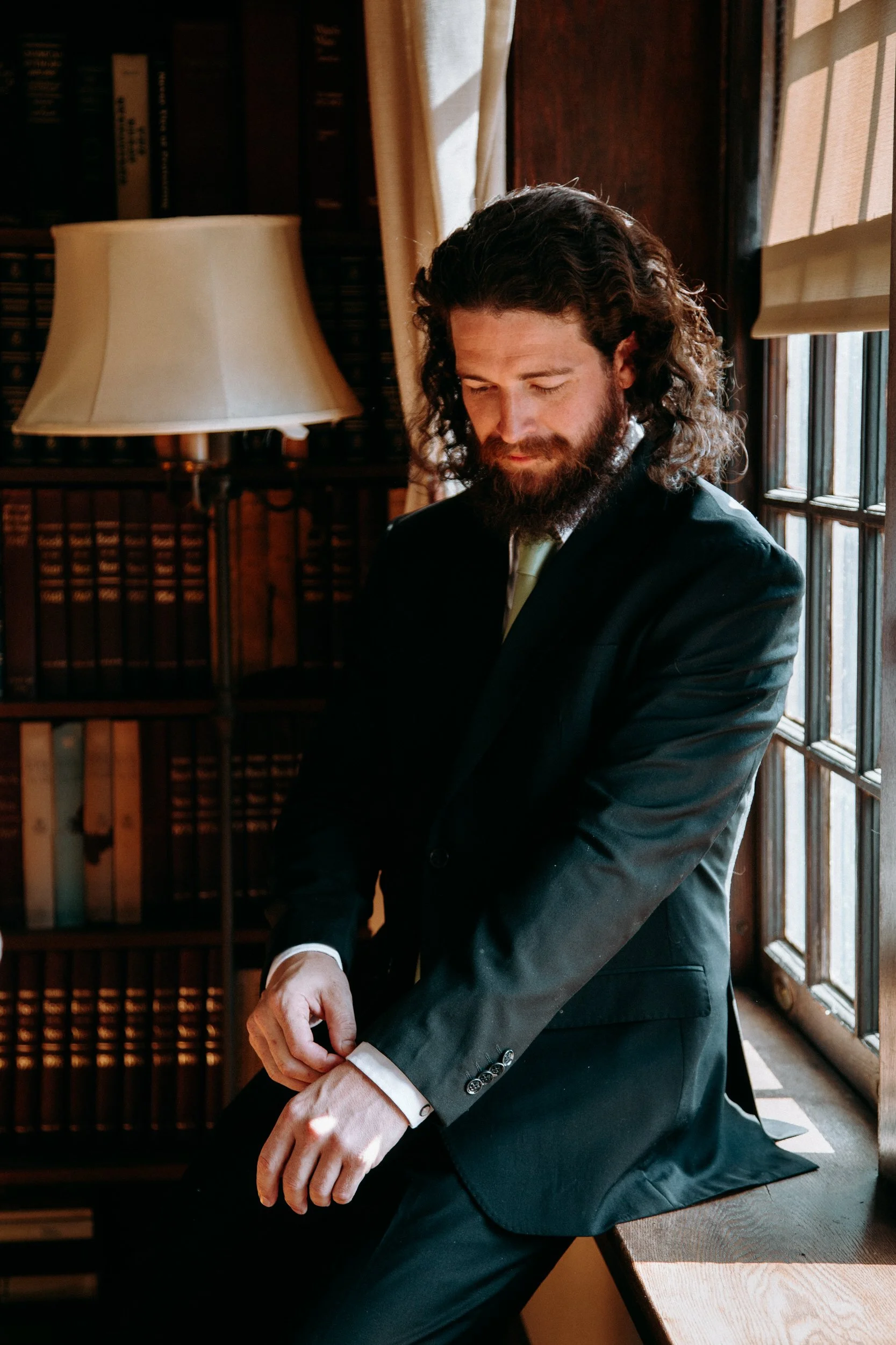 A man with long, curly hair and a beard, dressed in a black suit, is sitting by a window adjusting his cufflinks in a cozy, wood-paneled room filled with books and a lamp.