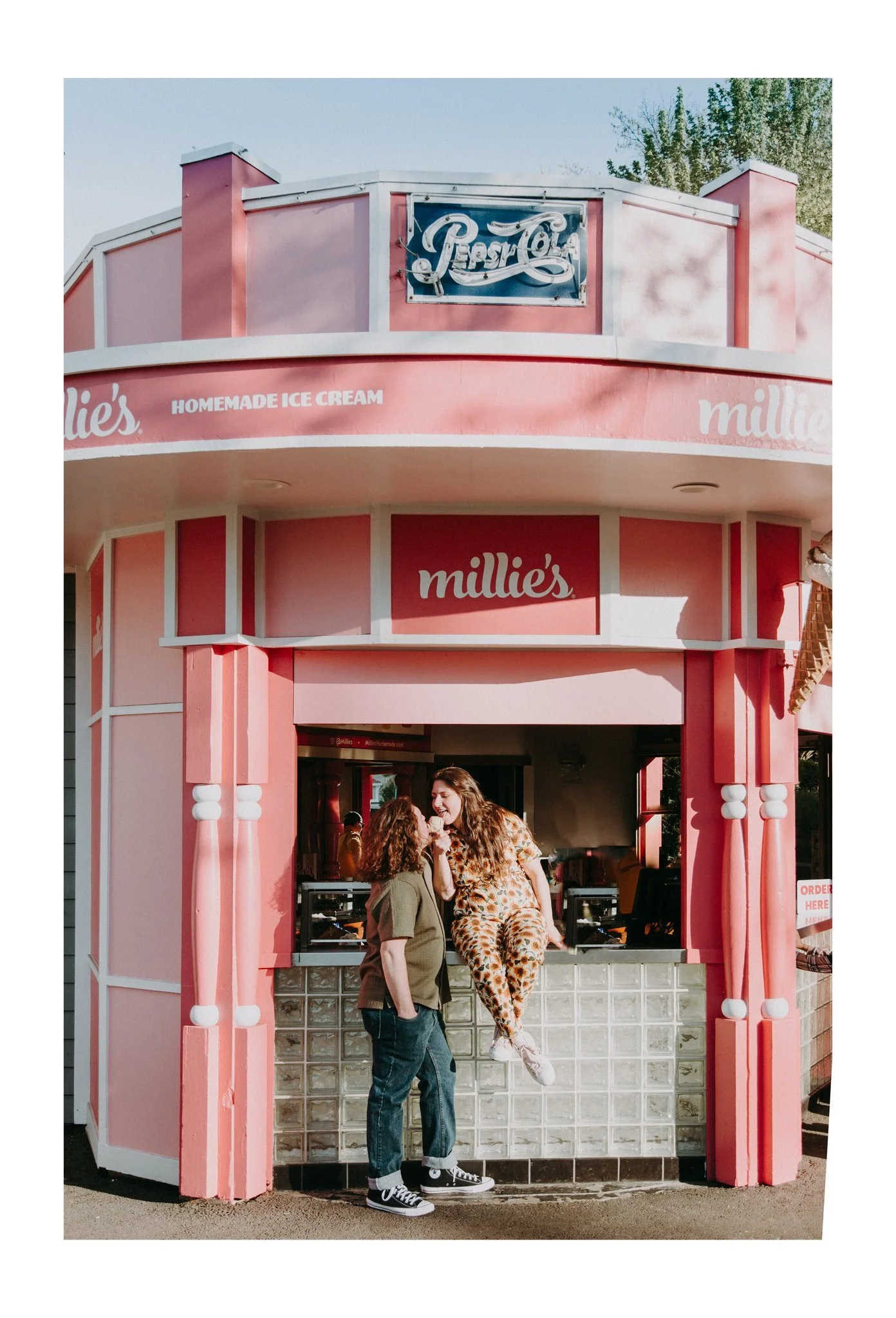 A couple leaning in to lick an ice cream cone as one sits on the counter of an outdoor ice cream shop