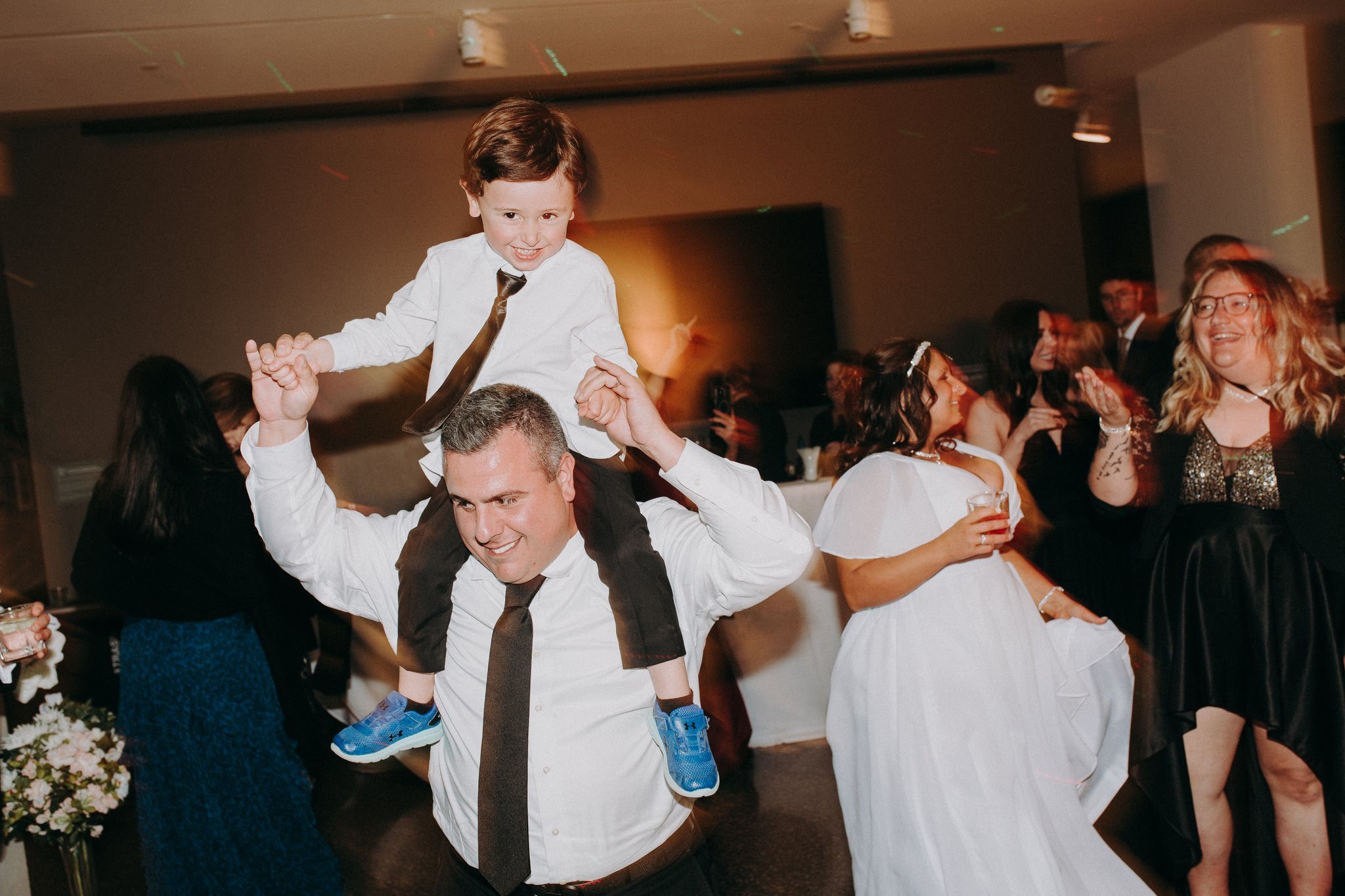 A child sitting on someone's shoulders as they dance at a wedding reception 