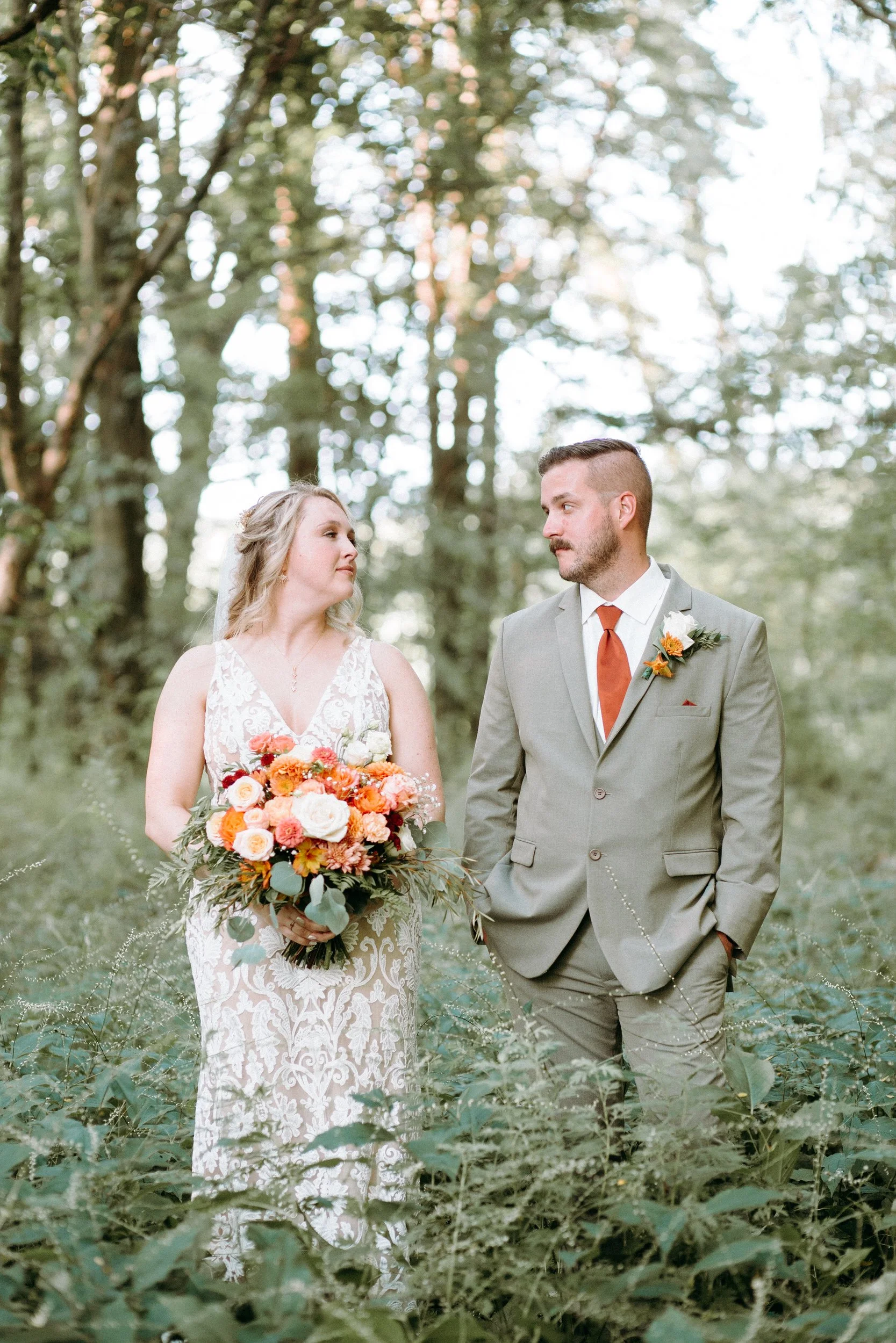 A newlywed couple standing side by side in a wooded area looking at each other