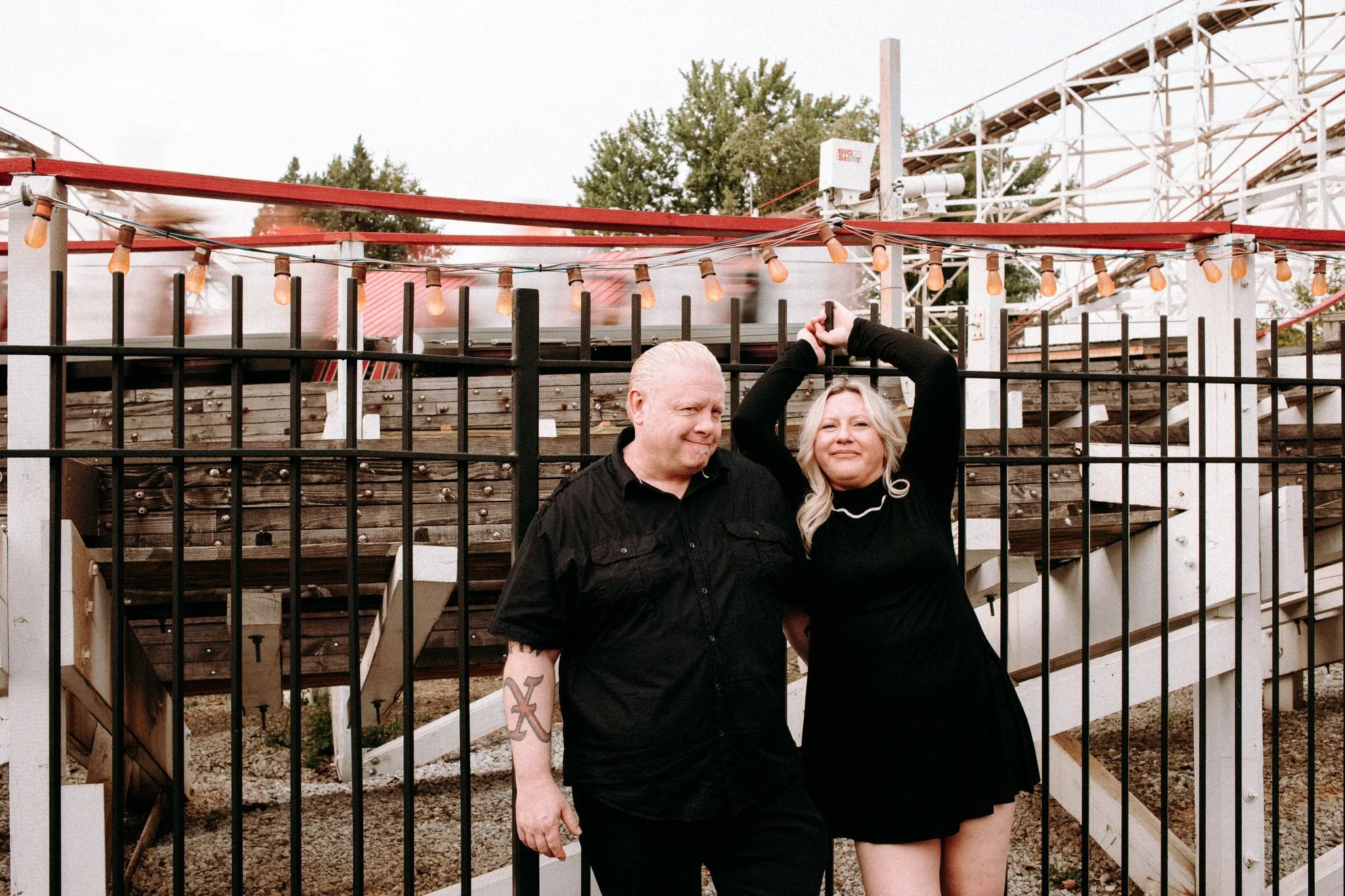 A couple leaning up against a metal fence in front of a roller coaster 