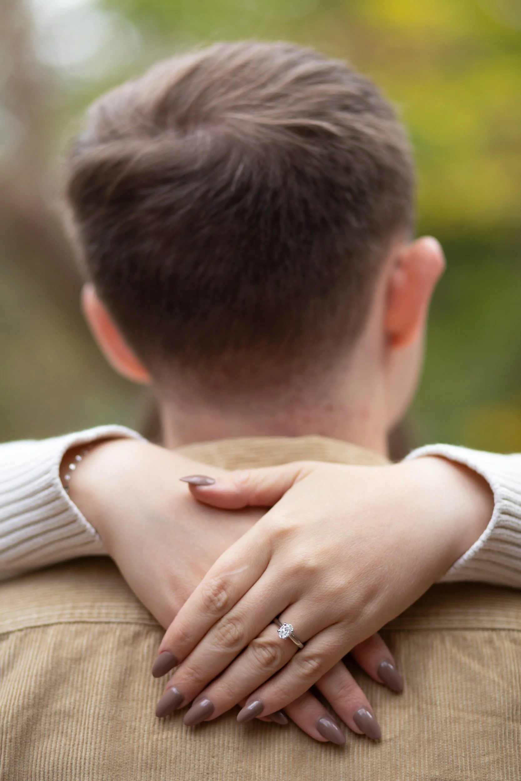 A person with their arms around their partner's shoulder showing off an engagement ring 