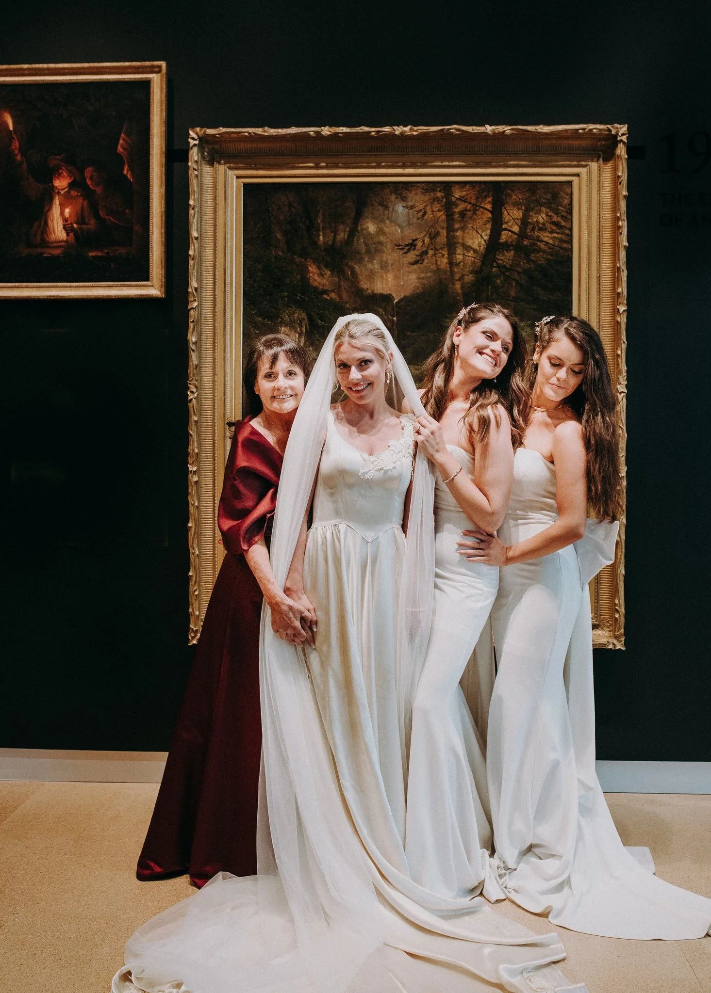 Four women in white wedding dresses and one woman in a dark red dress standing in front of a large framed painting with a forest scene, inside an art gallery.