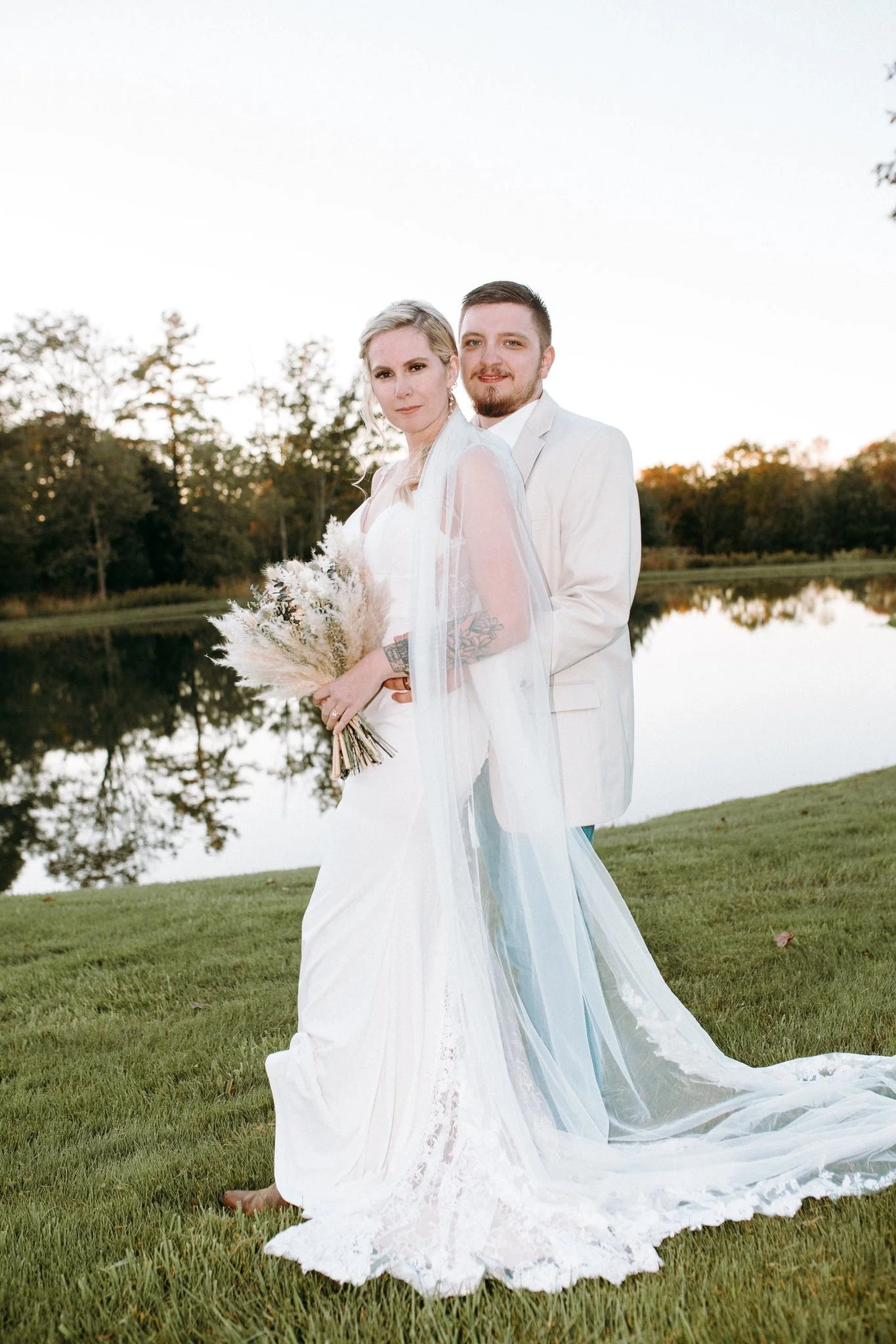 A newlywed couple standing one in front of the other as they stand next to a lake 