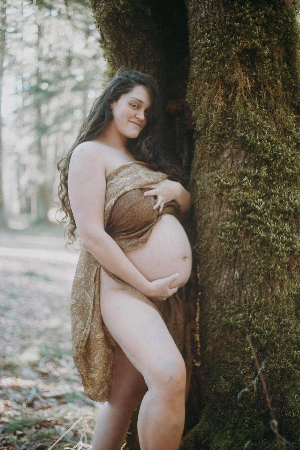 A pregnant person standing in a wooded area holding their stomach 