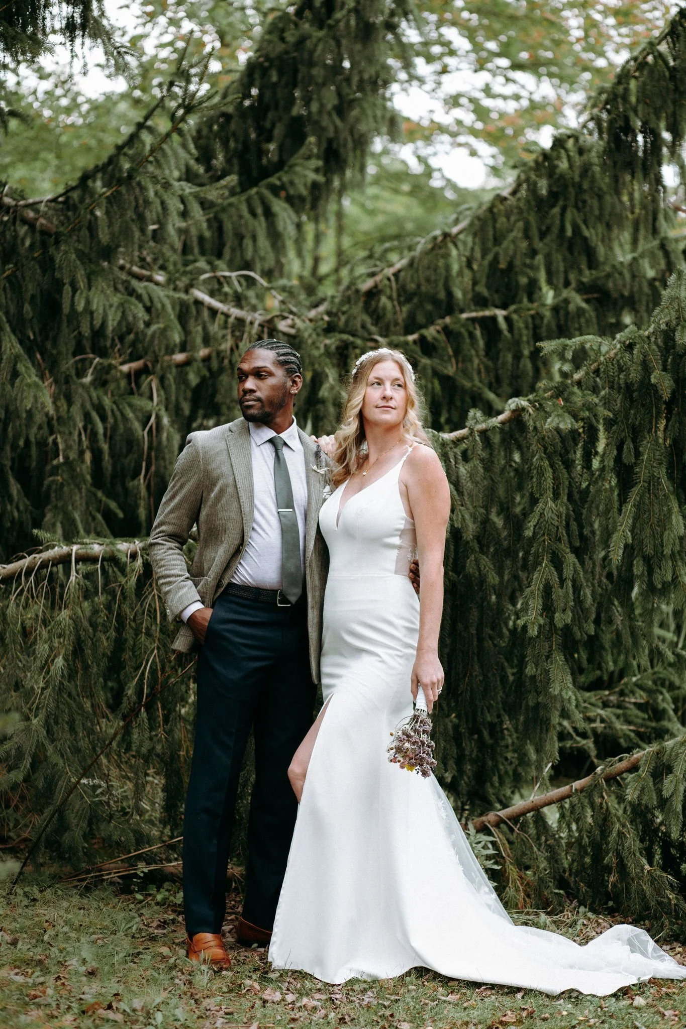 A bride and groom standing outdoors in front of a large pine tree. The groom has dark skin, braided hair, and is wearing a gray suit with a white shirt and dark tie. The bride has light skin and blonde hair, wearing a white wedding dress with a slit, holding a small bouquet of flowers. The setting looks like a forest or park.