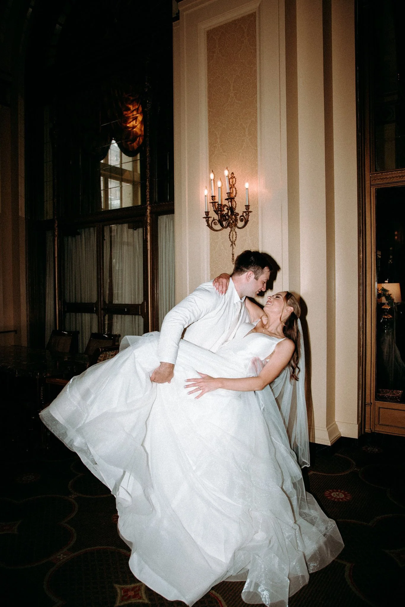 A couple in wedding attire dancing and smiling indoors near a wall with a chandelier and large window.