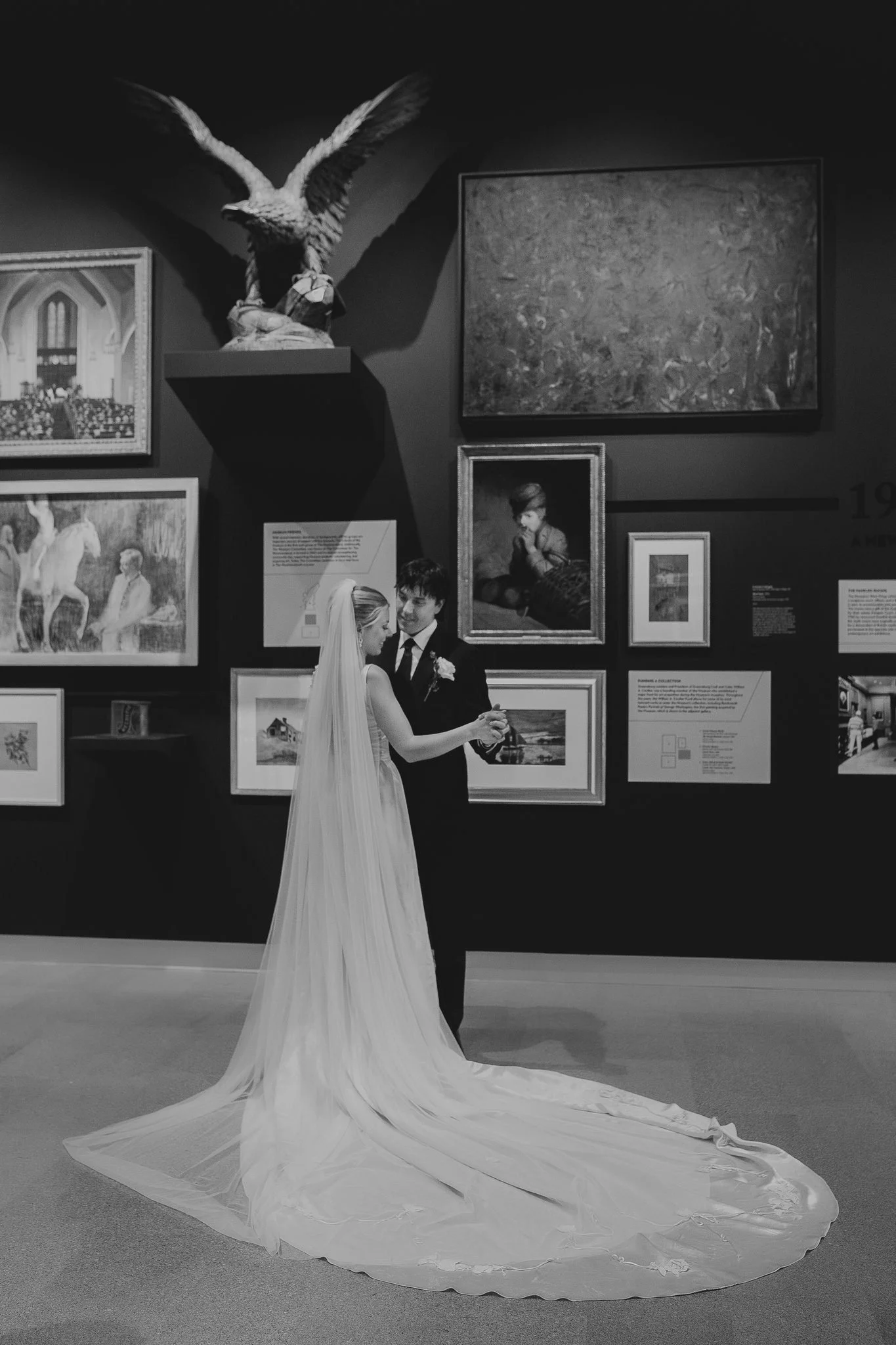 A bride and groom dance in an art gallery with framed paintings and a sculpture of an eagle in the background.