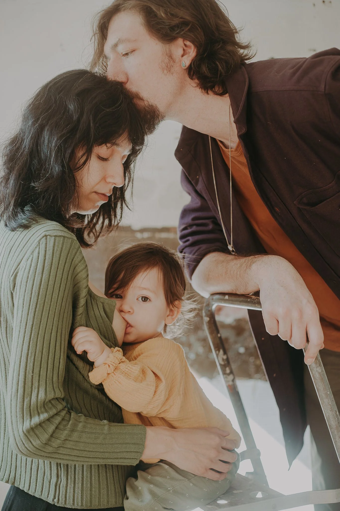 A person kissing their partner's head as she breast feeds their child 