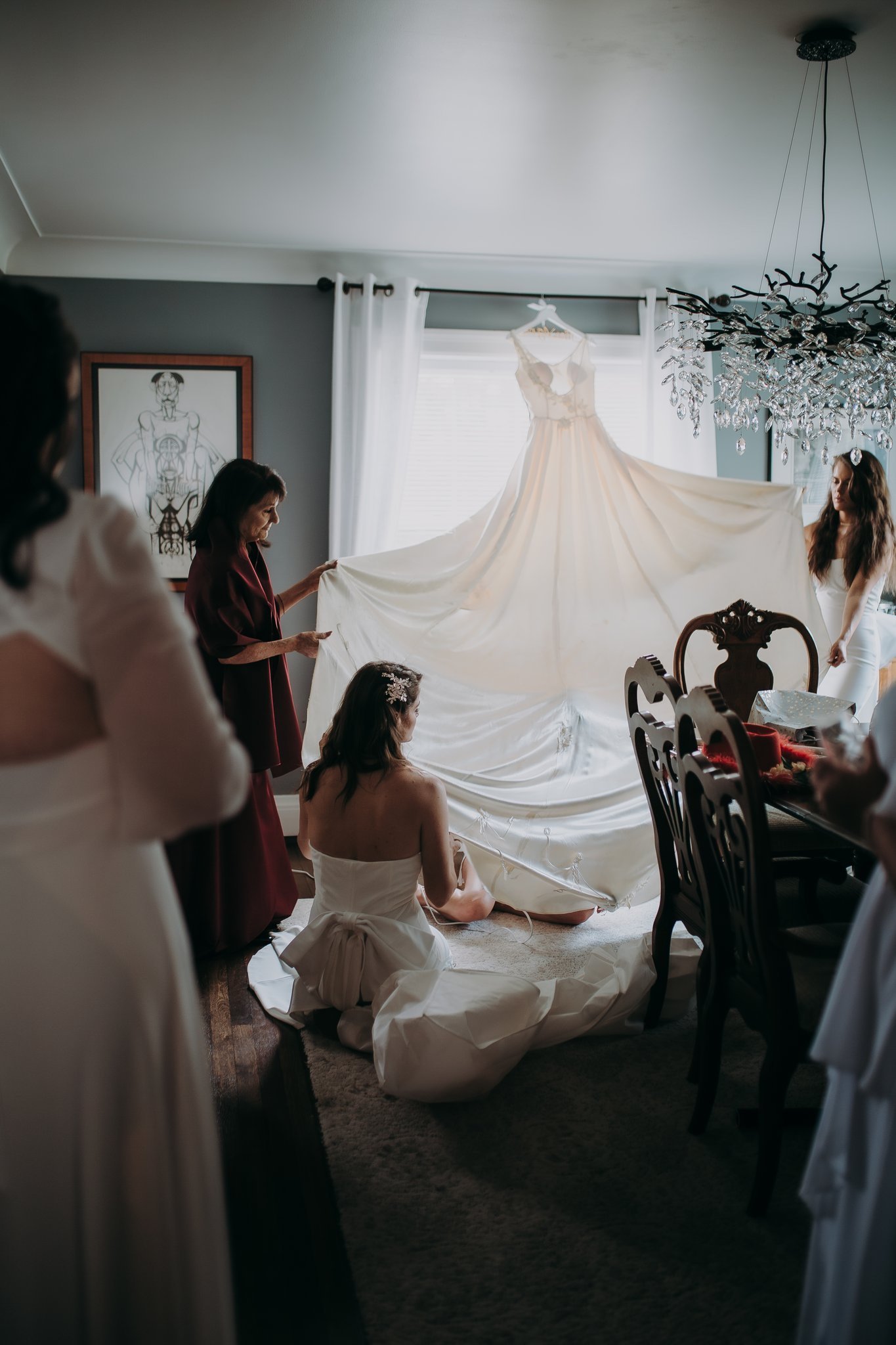 Two people holding up the bottom of a wedding dress hanging from a window