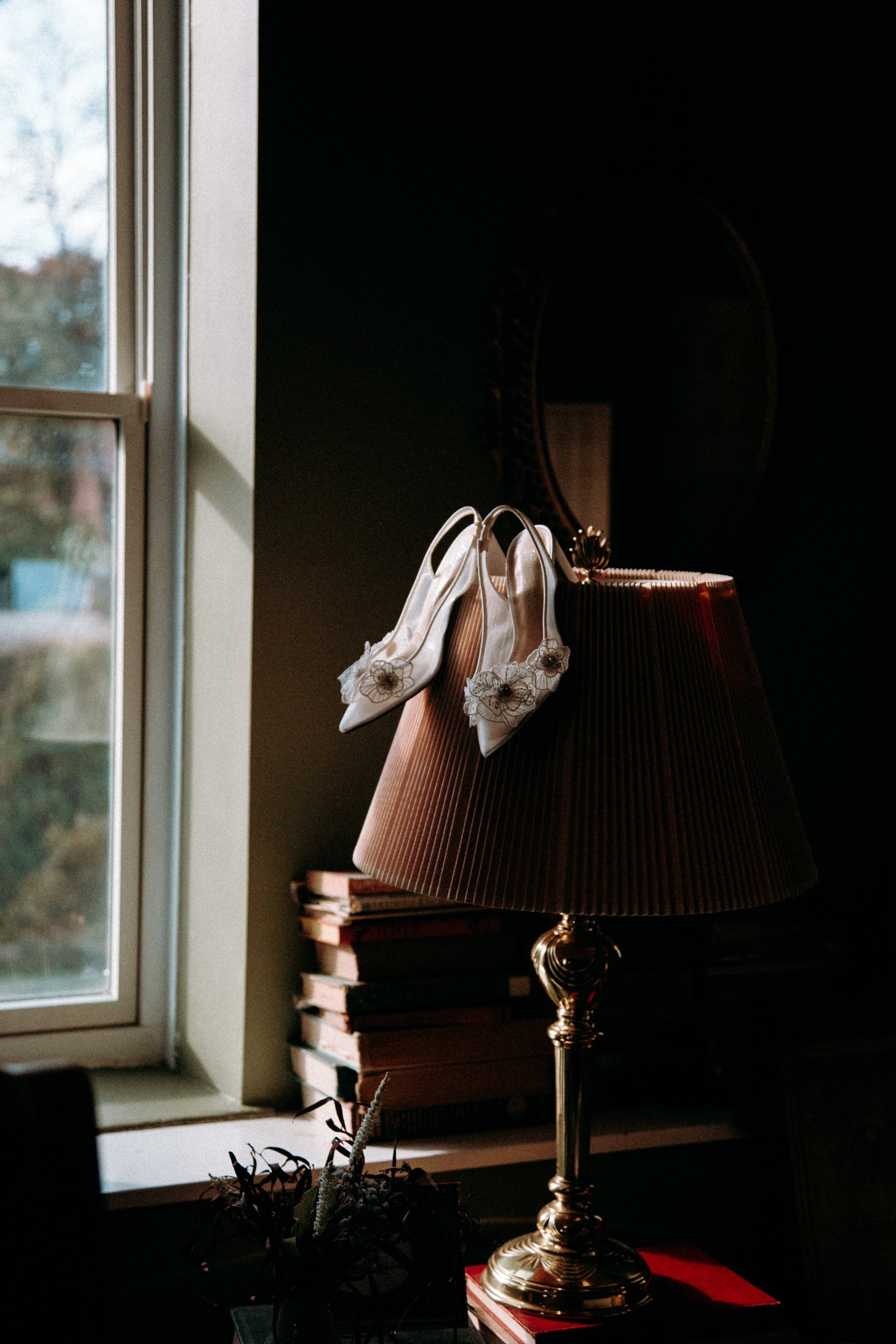 A pair of white shoes with floral embellishments hanging on a brass or gold-colored lamp near a window. A stack of books is on the windowsill, and a plant is in the foreground.