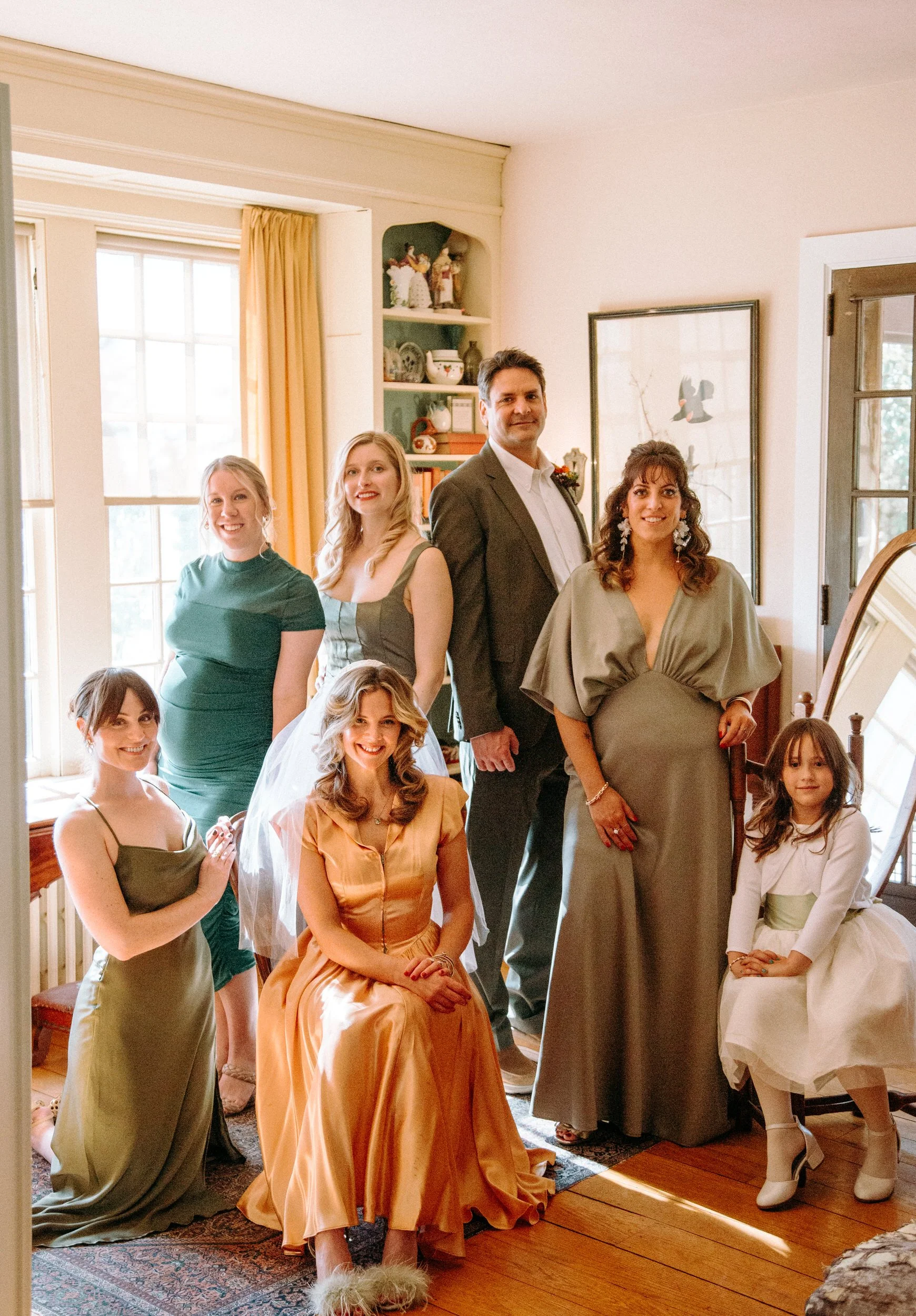 Group of seven people, including women, a man, and a young girl, posing indoors at a social gathering or celebration, in a room with large windows and decorative items.