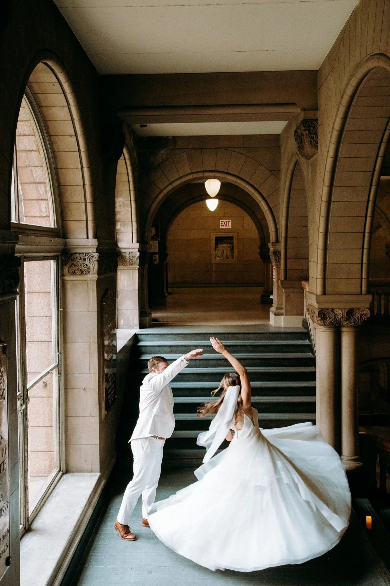 A bride and groom dancing inside a grand, arched hall with marble stairs and large windows, with the bride's white dress swirling around her as they dance.