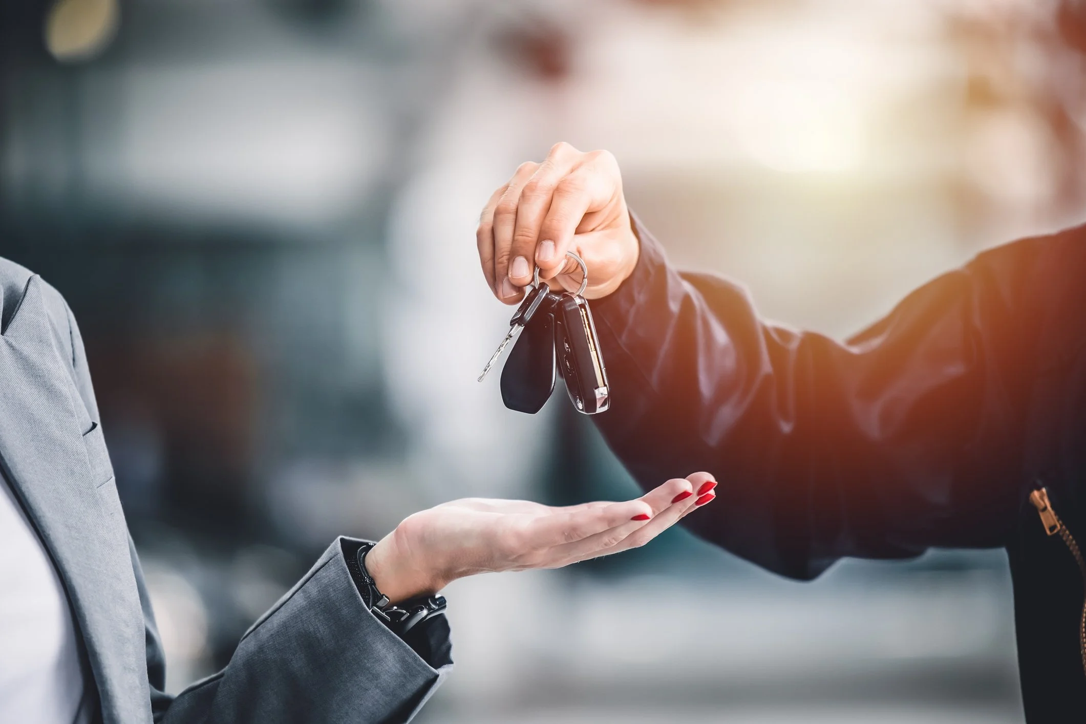 A person in a gray blazer is handing over keys to another person with red painted nails. The keys are being passed at the person's open hand.