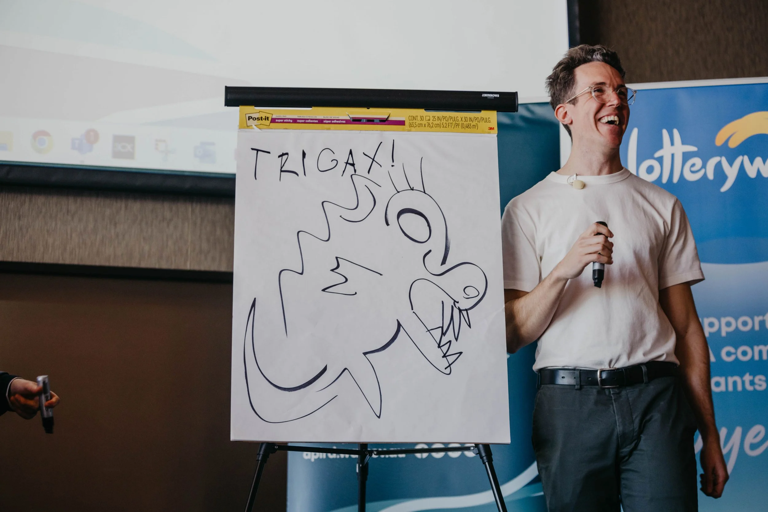 A man with glasses, holding a microphone, standing next to a whiteboard drawing of a dragon and the words "TRIGAX!" in a presentation room.