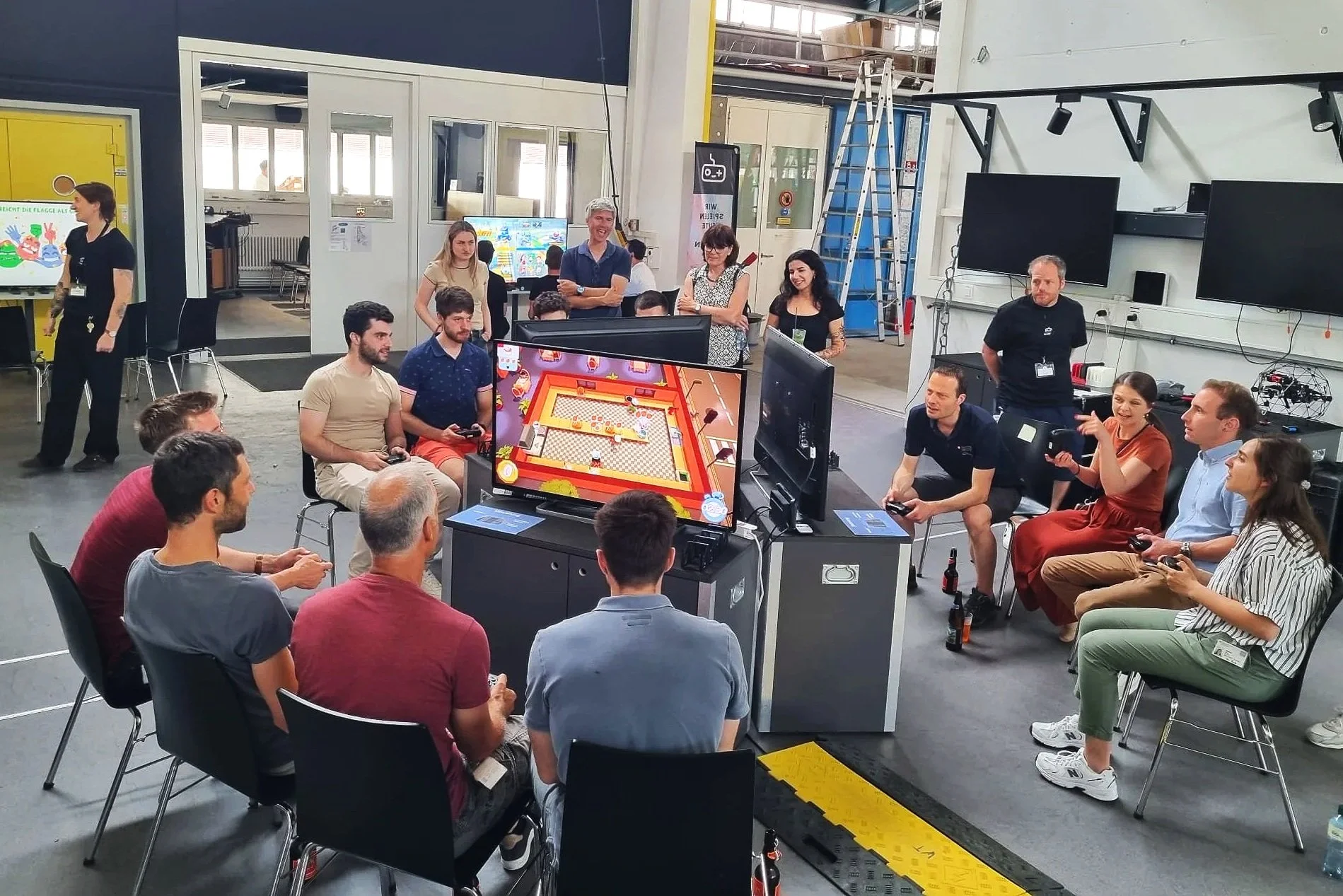 Menschen spielen ein Videospiel in einer Gruppe auf einer Veranstaltung, während andere zuschauen, in einem großen Raum mit Bildschirm und Technik.