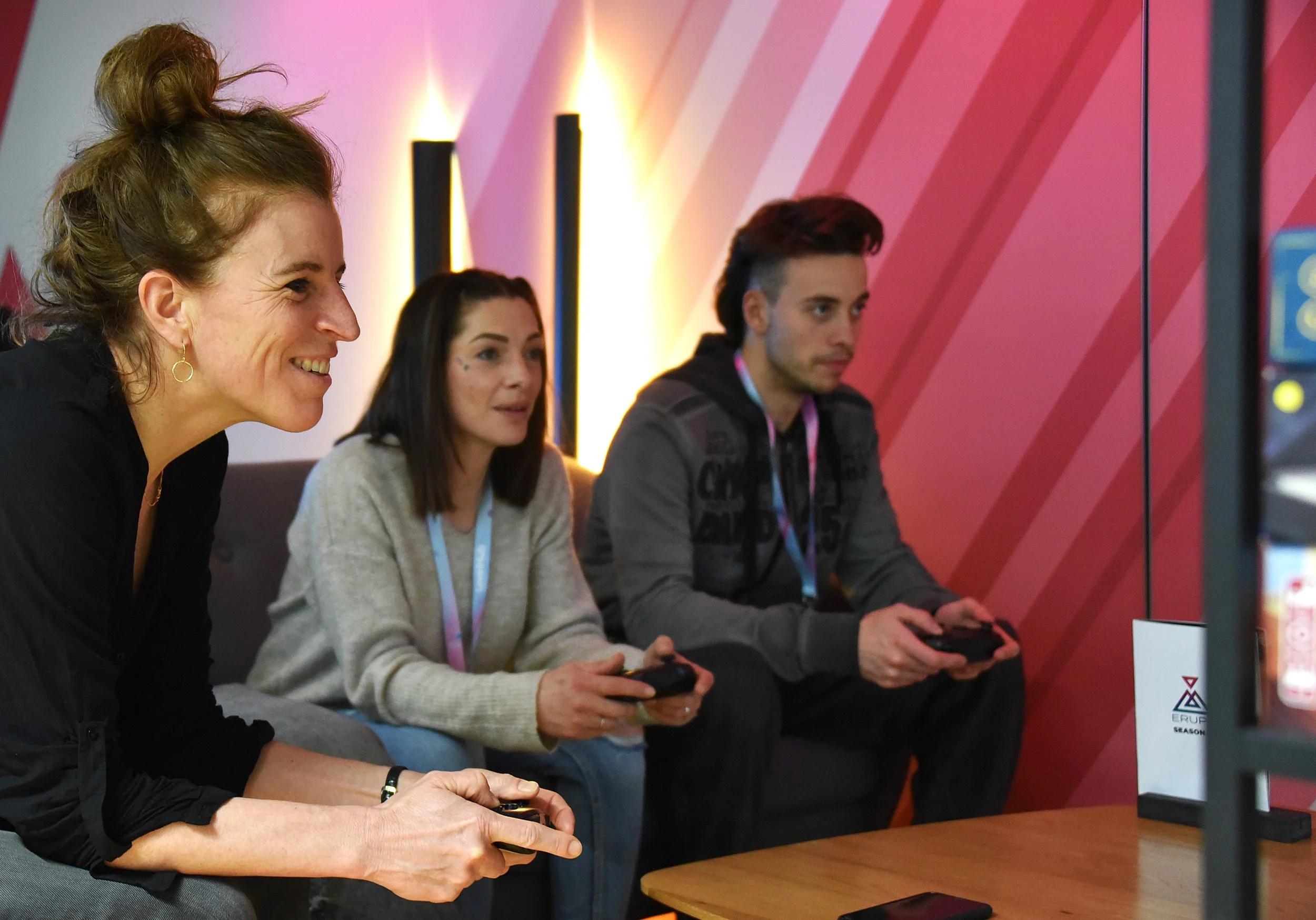 Drei Personen sitzen auf einem Sofa und spielen ein Videospiel, während eine Frau auf der linken Seite lacht, in einem Raum mit pink-gestreiften Wänden.