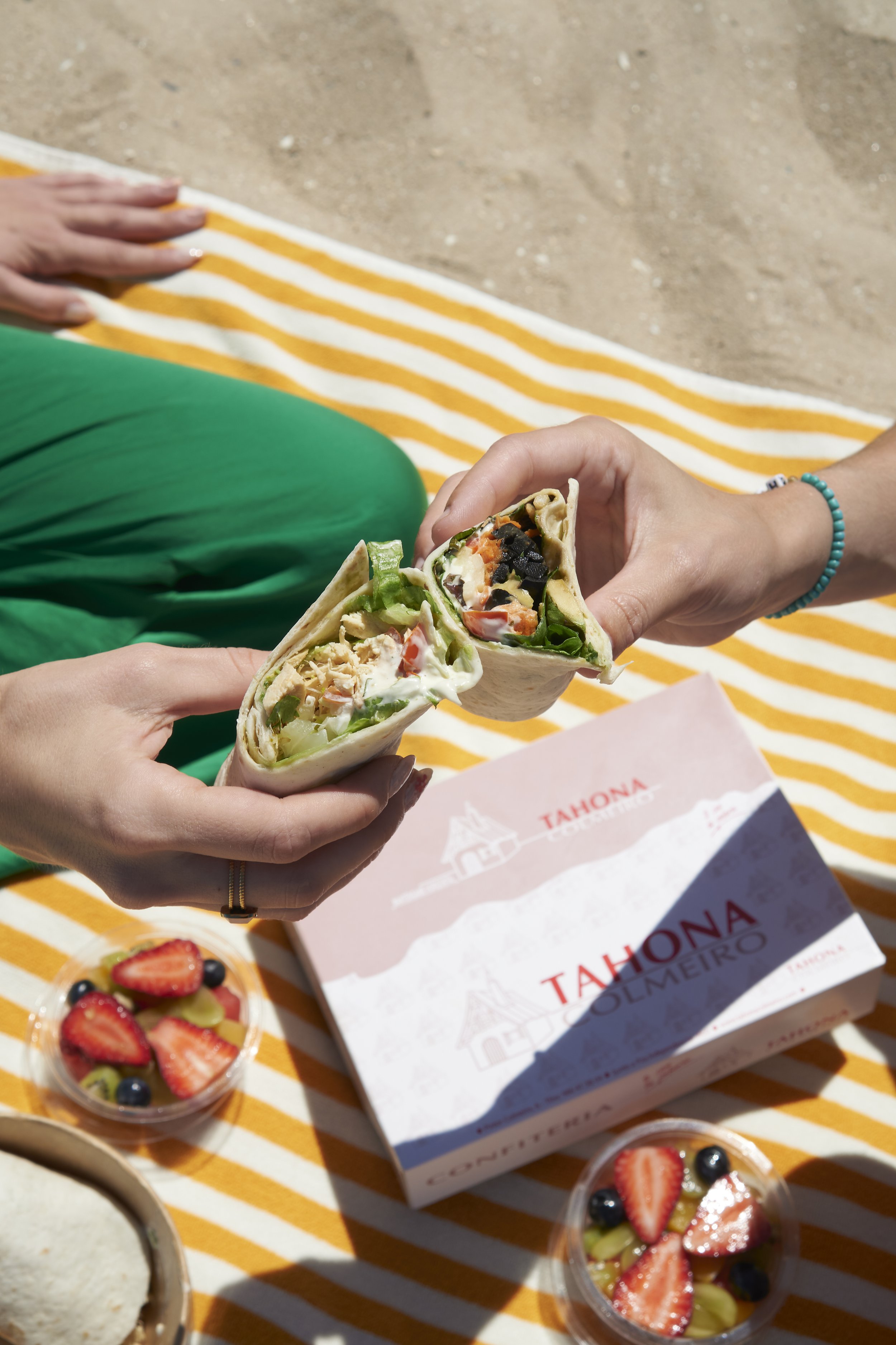 Dos personas sosteniendo un burrito cortado por la mitad, con un recipiente de fruta cortada a un lado y una caja de taquería en una manta a rayas amarillas y blancas.