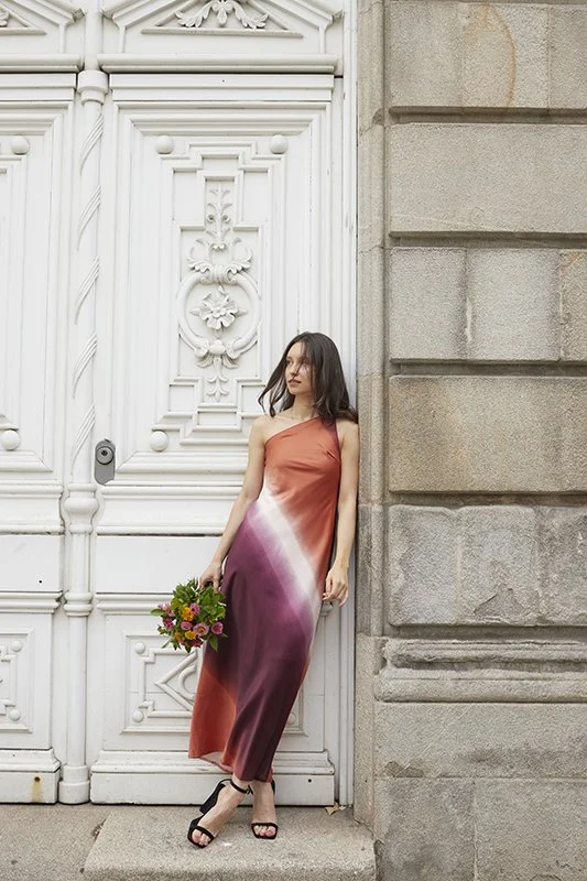 Una mujer con vestido largo de colores degradados en tonos naranja, rosa y morado, sosteniendo un ramo de flores, apoyada en una estructura de piedra y una puerta blanca decorada con detalles ornamentales.