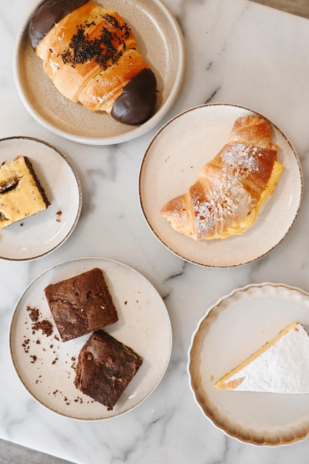 Variedad de postres en platos pequeños sobre una mesa de mármol, incluyendo croissant relleno, brownies, y otros dulces.
