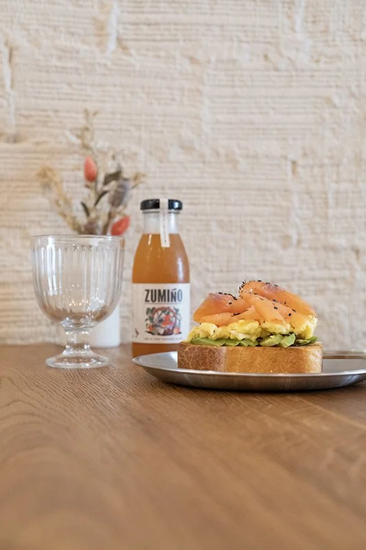 Una rebanada de pan con aguacate, salmón y pimienta en un plato, acompañado de una botella de jugo y un vaso vacío en una mesa de madera.