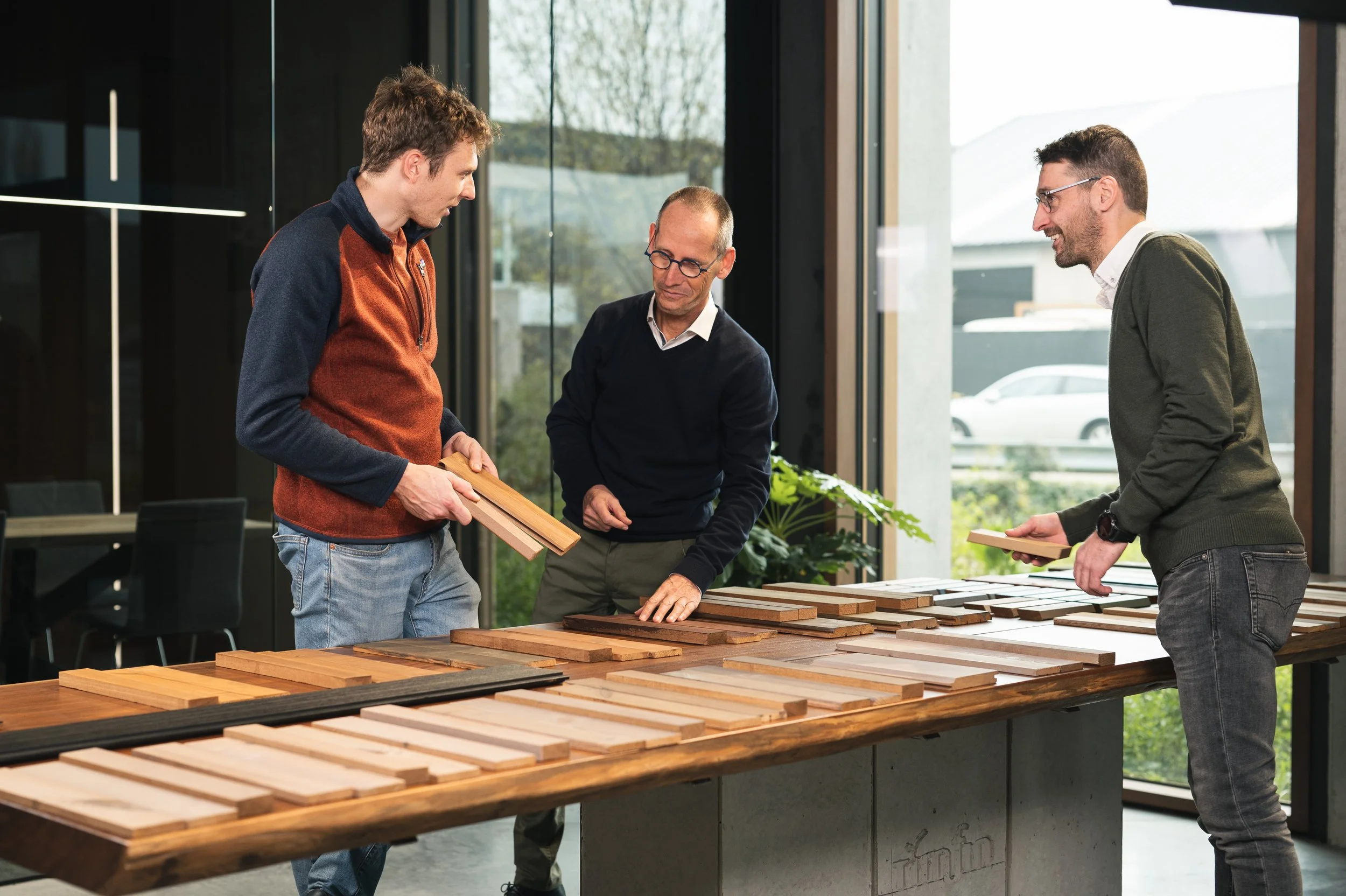 Drie mannen bekijken houten planken en materiaal op een tafel in een showroom of werkruimte met grote ramen.
