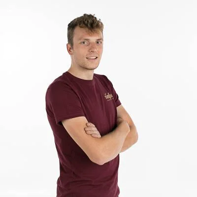 Een jonge man met kort, lichtbruin haar draagt een maroon T-shirt en staat met gekruiste armen tegen een witte achtergrond.