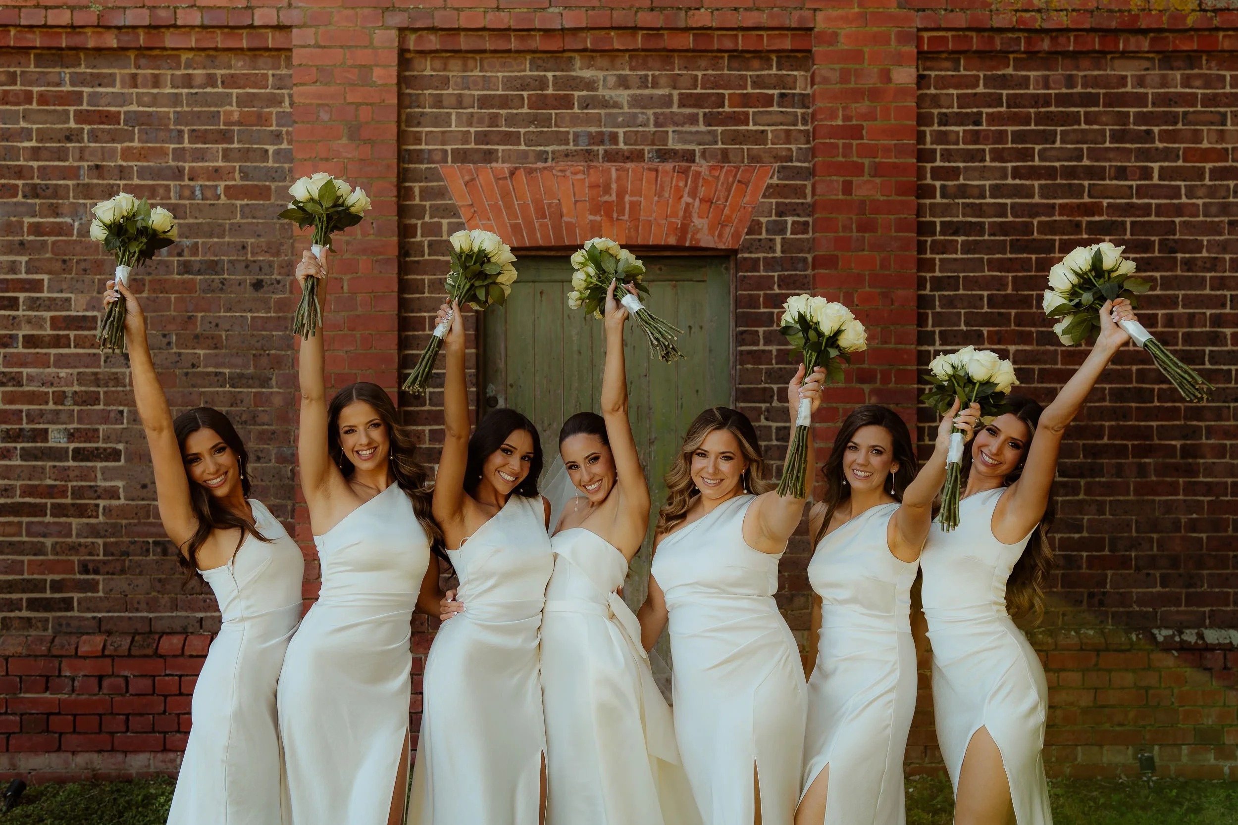 7 bride and bridesmaids pumping their matching white rose bouquets above their heads in matching white dresses in front of the stables at Stones of the Yarra Valley