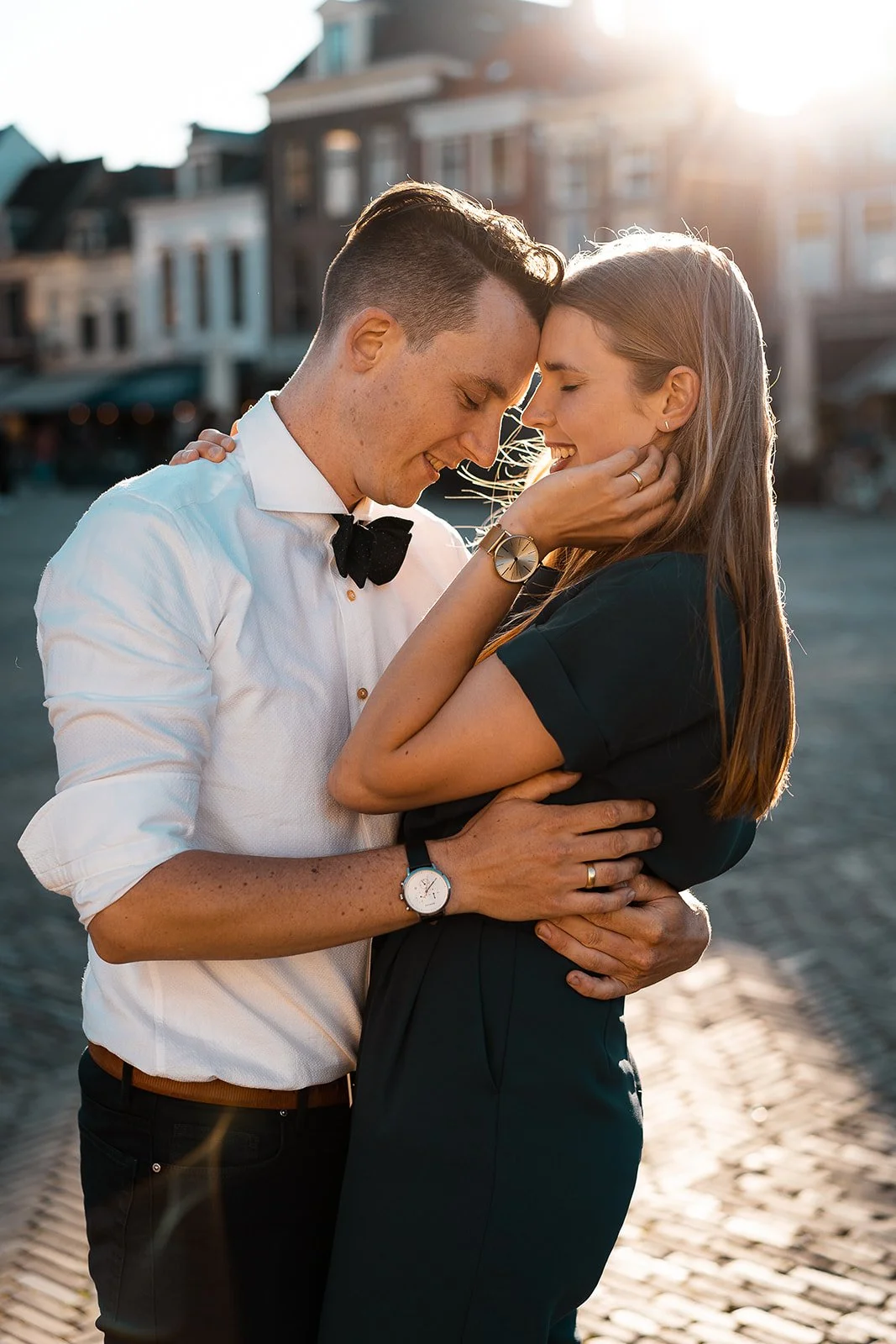 A young couple embracing on a sunny street with the old buildings of Delft in the background, smiling with foreheads touching.