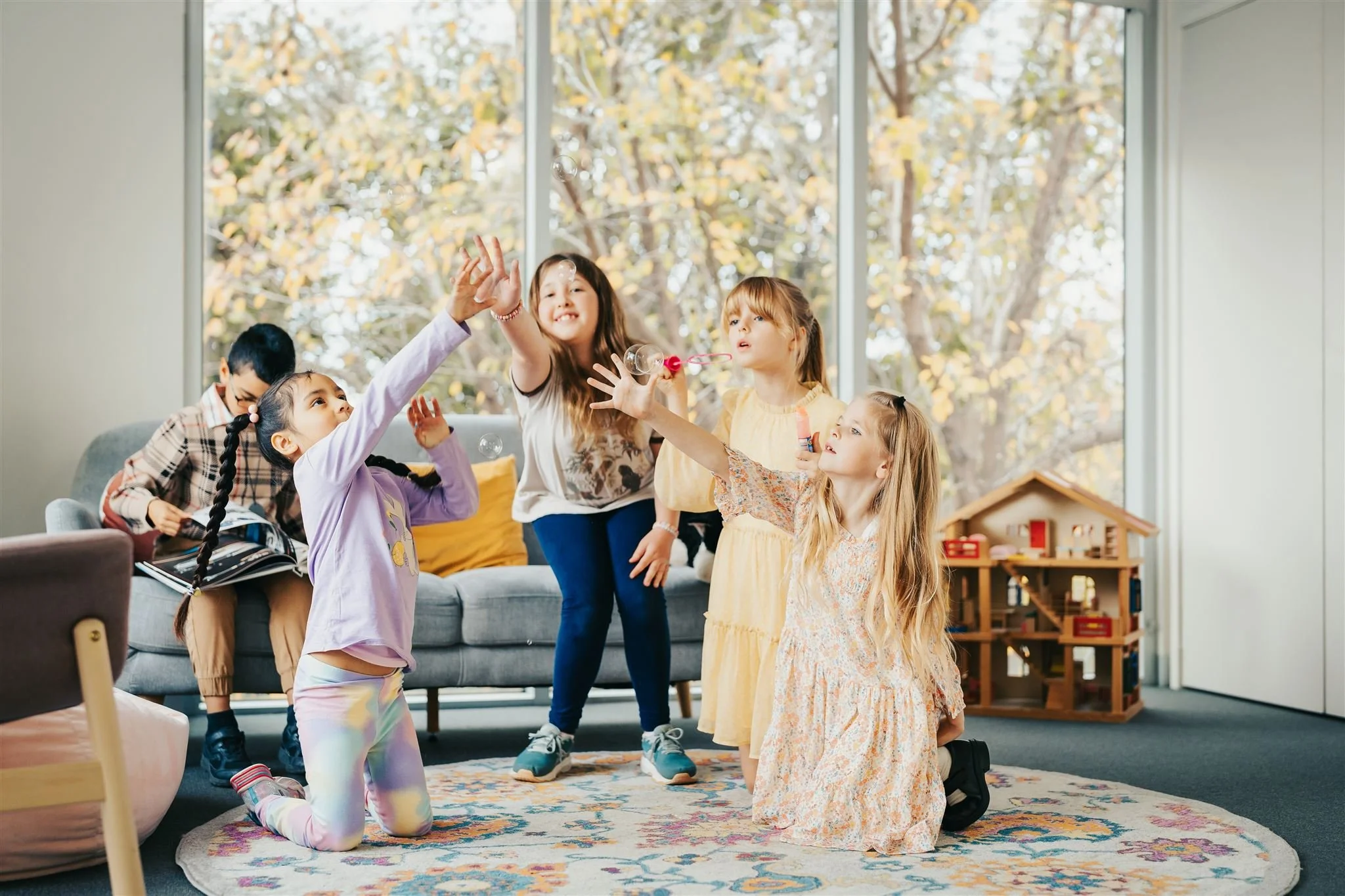 Commercial photo of five children playing with soap bubbles in a living room with a large window showing autumn trees outside. One girl is kneeling and reaching for bubbles, while others stand or kneel, smiling and interacting