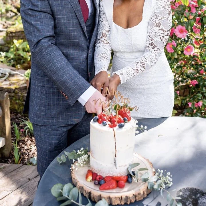 A bride and groom standing side by side, cutting a wedding cake outdoors surrounded by greenery and pink flowers. The cake is decorated with berries and has a gold topper.