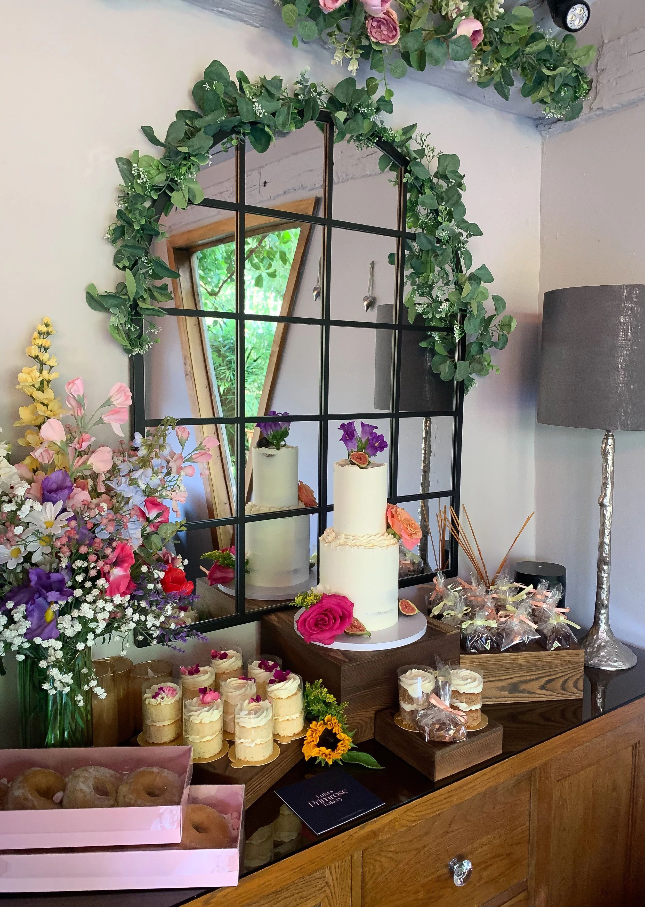 A display of baked goods and flowers at a celebration or bakery. There is a two-tier white cake with pink roses and figs as decoration, surrounded by a variety of other pastries, cupcakes, and decorative flowers. Behind the display, a mirror and a window with greenery outside are visible.