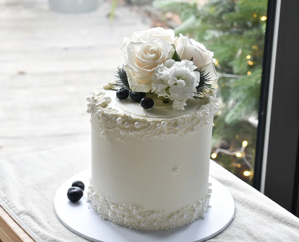 A white iced cake decorated with white roses, white flowers, black berries, and small pearl-like decorations on top and around the edges, sitting on a white cake board on a table near a window.